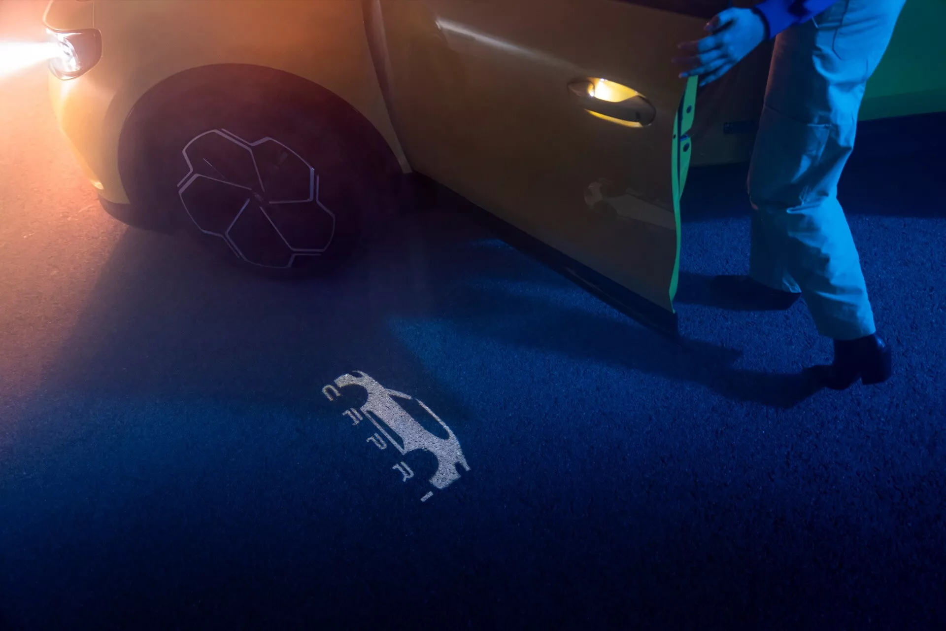 Częściowy widok Forda Capri 2024 z niskiego kąta, skupiający się na boku w pobliżu przednich drzwi, pokazujący dolną część przednich drzwi z nowoczesną, zintegrowaną klamką i otwartymi drzwiami. Widoczny jest symbol świetlny rzucany na ziemię przez technologię welcome/puddle light, oświetlający niebieskie i pomarańczowe odcienie w słabo oświetlonym nocnym środowisku. Przednie koło i opona są częściowo widoczne, z dodatkowym blaskiem jasnego źródła światła w prawym dolnym rogu.
