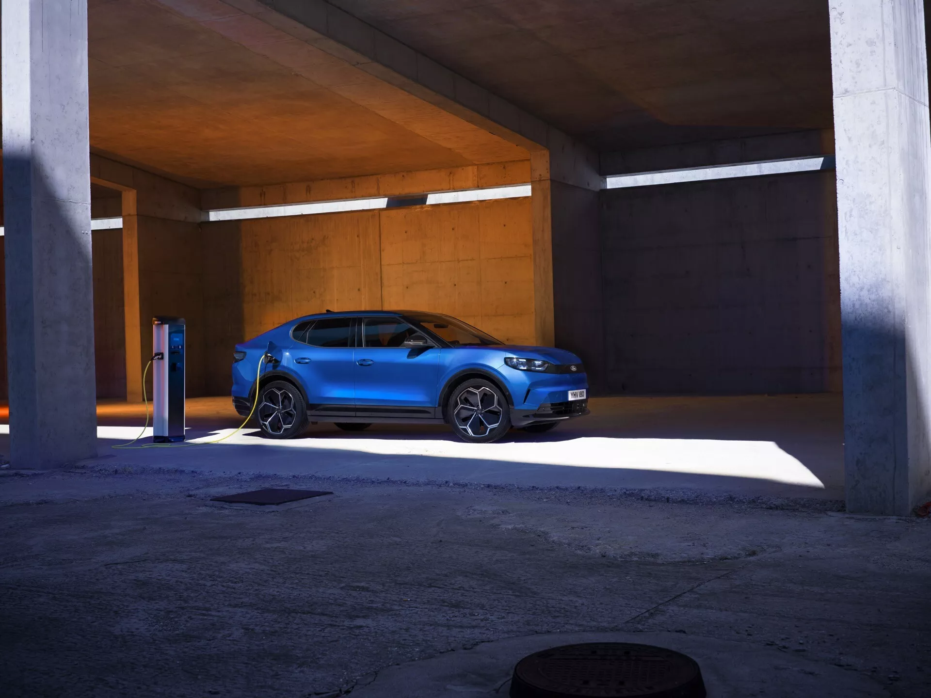 Jasno niebieski SUV Ford Capri 2024 zaparkowany w betonowej strukturze parkingowej, przedstawiony z przedniego widoku trzy-czwarte, ukazujący swoje kątowe reflektory LED, charakterystyczny design kraty i podłączoną stację ładowania pojazdów elektrycznych, podkreślając jego nowoczesny, sportowy design oraz zdolności elektryczne.