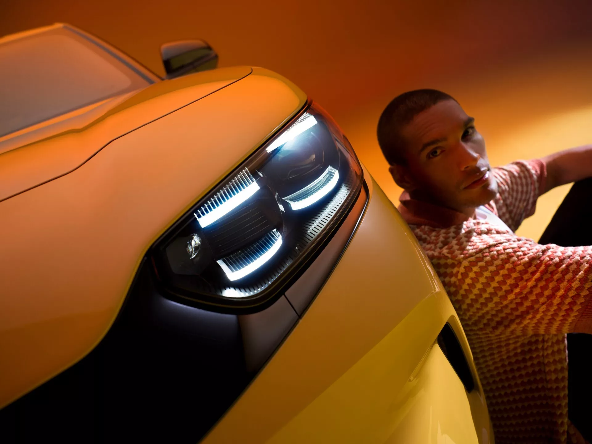 Zbliżenie lewego przedniego kwartału Forda Capri 2024, z naciskiem na zespół reflektorów LED z wieloma elementami, w tym światłem dziennym w kształcie litery 'L'. Jaskrawo żółty samochód jest zrobiony pod niskim kątem, dominując nad ciemniejszym tłem. Osoba opiera się o samochód, patrząc poważnie w obiektyw, dodając do kompozycji element stylu życia, układ jest nowoczesny i wyszukany.