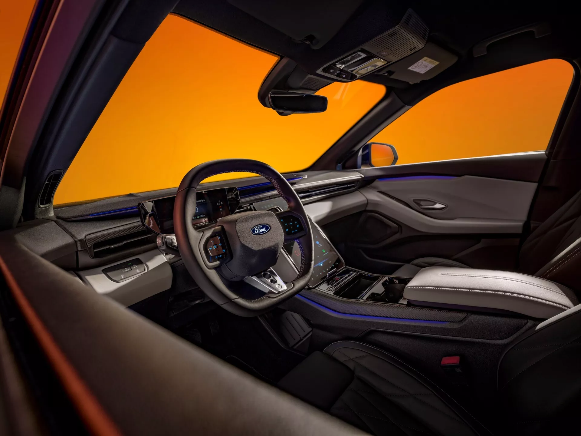 Wnętrze Forda Capri 2024 czwartej generacji, widok z perspektywy tylnego pasażera. Wyeksponowana kierownica z logo Ford oraz zamontowane kontrolki do sterowania dźwiękiem, tempomatem, komendami głosowymi i obsługą telefonu bez użycia rąk. Widzoczny jest cyfrowy zestaw wskaźników z prędkościomierzem, obrotomierzem i innymi wskaźnikami oraz centralny ekran informacyjno-rozrywkowy prawdopodobnie oferujący opcje nawigacji i media. Wnętrze pokazuje ekskluzywne połączenie skórzanych siedzeń z kontrastującymi szwami, metalowych i błyszczących czarnych powierzchni. Funkcje bezpieczeństwa obejmują poduszki powietrzne, pasy bezpieczeństwa i zaawansowane systemy wspomagania kierowcy. Dwukolorowy schemat wnętrza podkreśla nowoczesną i luksusową estetykę, wyróżnioną w promocyjnym zdjęciu z pomarańczowym tłem.