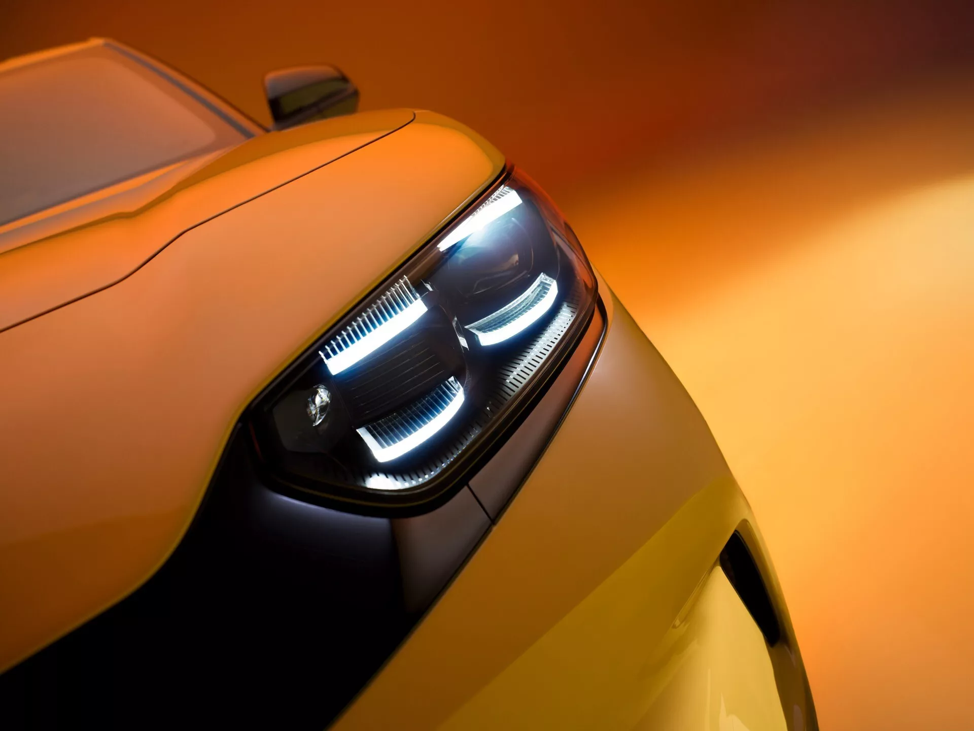 Zbliżony, ukośny widok przedniego lewego boku Forda Capri 2024, skupiający się na skomplikowanym designie reflektora z kątowymi i warstwowymi światłami do jazdy dziennej oraz głównym reflektorem. Obudowa reflektora jest szczelna, z przezroczystą osłoną z poliwęglanu. Pojazd, pomalowany na ciepły odcień żółtego, odbija światło, nadając luksusowy i zaawansowany technologicznie wygląd. Obraz ma małą głębię ostrości, podkreślając reflektor, podczas gdy tło pozostaje delikatnie rozmyte.