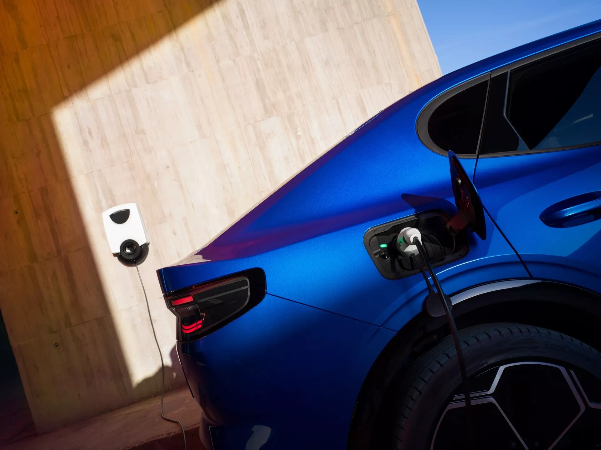 Częściowy widok niebieskiego Forda Capri 2024 (4. generacja) ładującego się na stacji ładowania pojazdów elektrycznych, uchwycony pod tylnym kątem trzech czwartych z naciskiem na prawą tylną część, pokazujący charakterystyczny design tylnych lamp LED, skomplikowane felgi wieloramienne i otwarty port ładowania podłączony do kabla. Obraz podkreśla sportowe krzywizny i dynamiczne linie samochodu przy jasnym świetle dziennym, z rozmytym tłem dzięki płytkiej głębi ostrości.