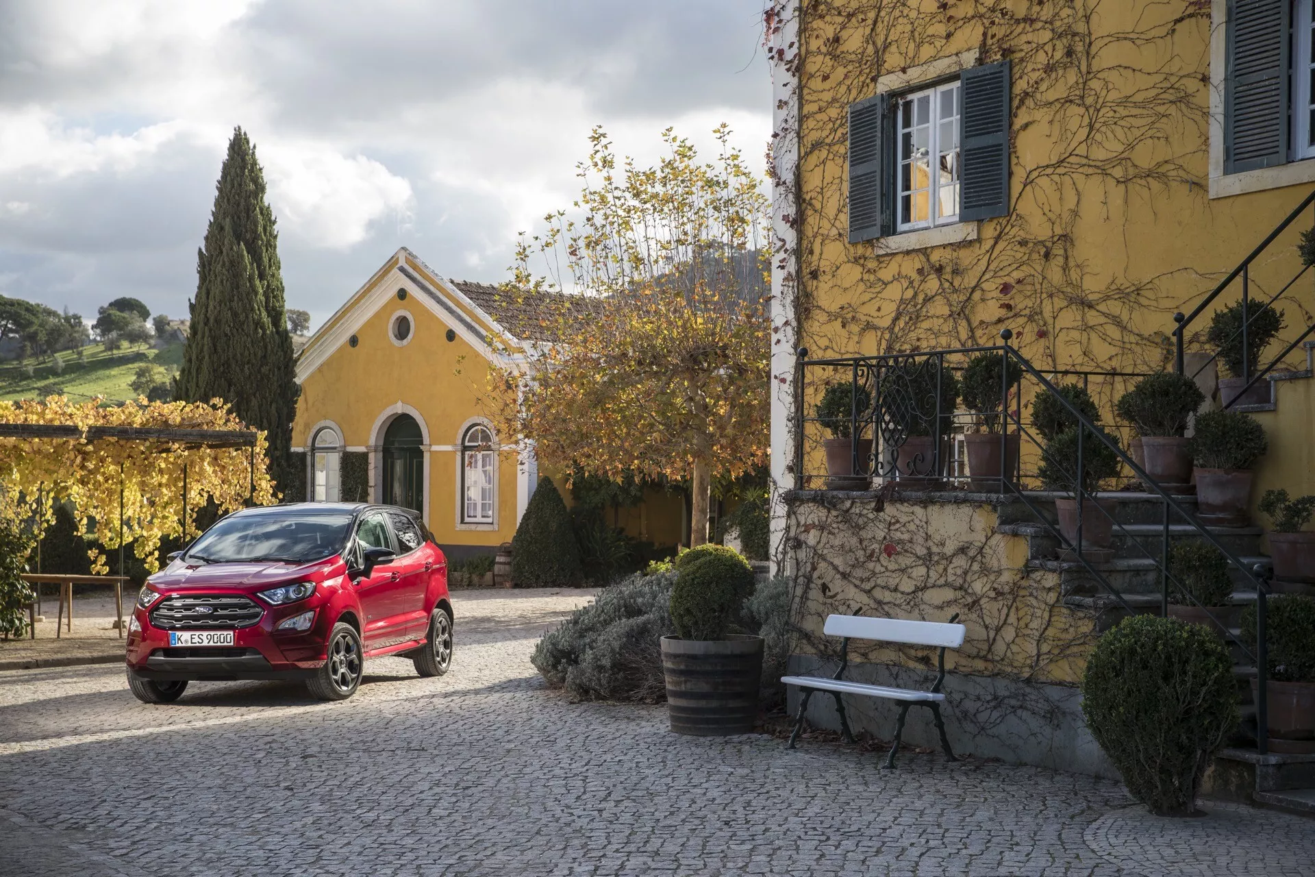 Ford EcoSport 2017 (1. generacja po face liftingu) w kolorze czerwonym, zaparkowany przed ochrowym domem z pnącymi roślinami, pod miękkim światłem porannym lub popołudniowym, prezentujący przedni i boczny profil pojazdu, stylowe felgi aluminiowe i nowoczesny design reflektorów LED