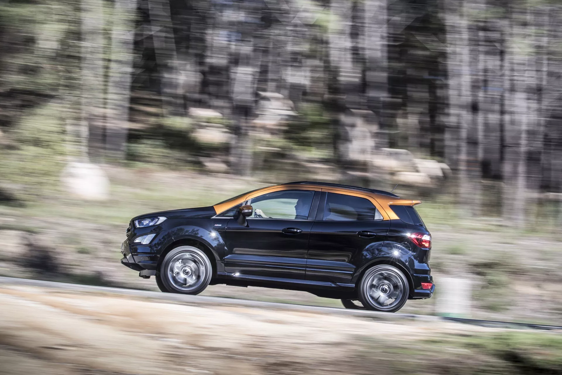 Ford EcoSport 2017 pierwszej generacji po liftingu w dynamicznym ruchu, podkreślając dwukolorową czarną i możliwą pomarańczową lub brązową kolorystykę, boczne i tylne cechy wzornictwa, felgi aluminiowe i połysk na drodze gruntowej lub żwirowej, sugerując zdolność jazdy w terenie.
