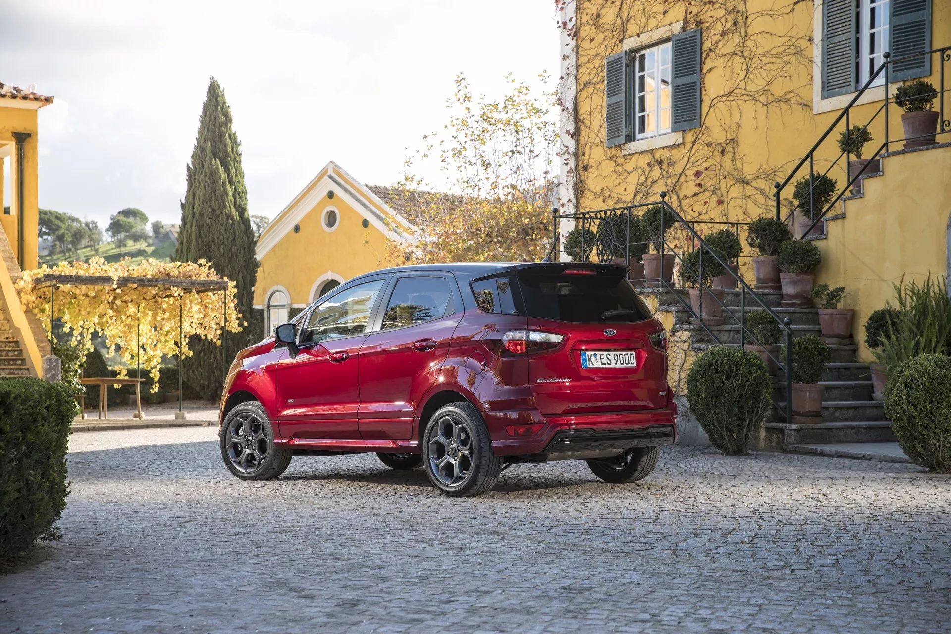 Ford EcoSport z 2017 roku, 1. generacja po liftingu, w kolorze czerwonym, prezentujący tył pojazdu z perspektywy trzech czwartych zaparkowany na nawierzchni z kostki brukowej z widocznymi szczegółami takimi jak LEDowe światła tylne, felgi ze stopów lekkich, zamazana tablica rejestracyjna w stylu europejskim, spoiler dachowy oraz czarne obramowania łuków kół na tle spokojnego otoczenia mieszkalnego z klasyczną architekturą i bujną roślinnością pod naturalnym światłem słonecznym.