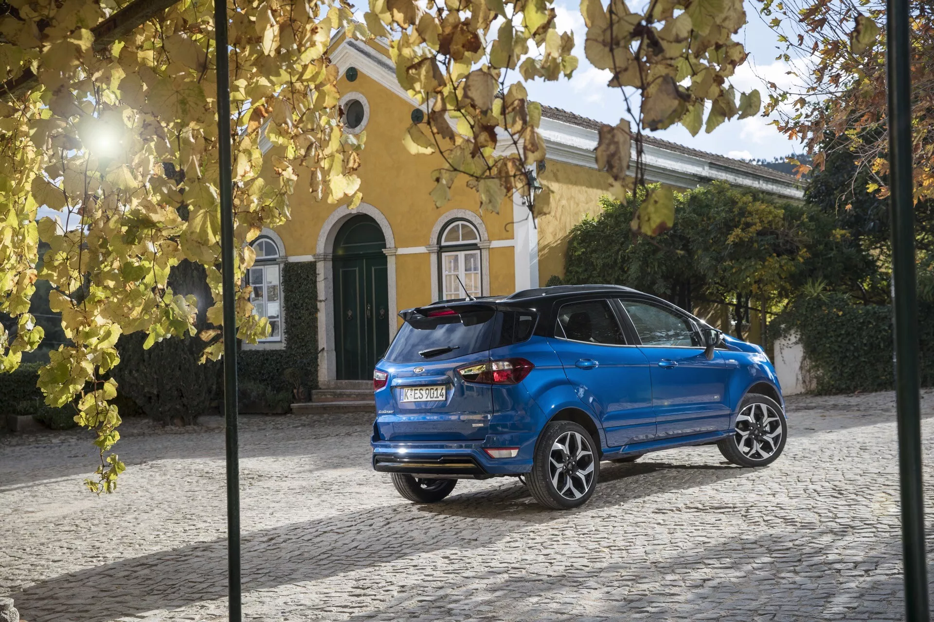 Tylny trzy czwarte widok Forda EcoSport (1 generacja - lifting) 2017 w kolorze niebieskim, przedstawiający tylne światła LED, spoiler z tyłu, szybę z tyłu z funkcją rozmrażania, wycieraczkę tylną, antenę w kształcie płetwy rekina, czujniki parkowania, podwójny układ wydechowy i felgi aluminiowe wieloszprychowe, zaparkowany na bruku obok żółtej willi z zielonymi okiennicami.