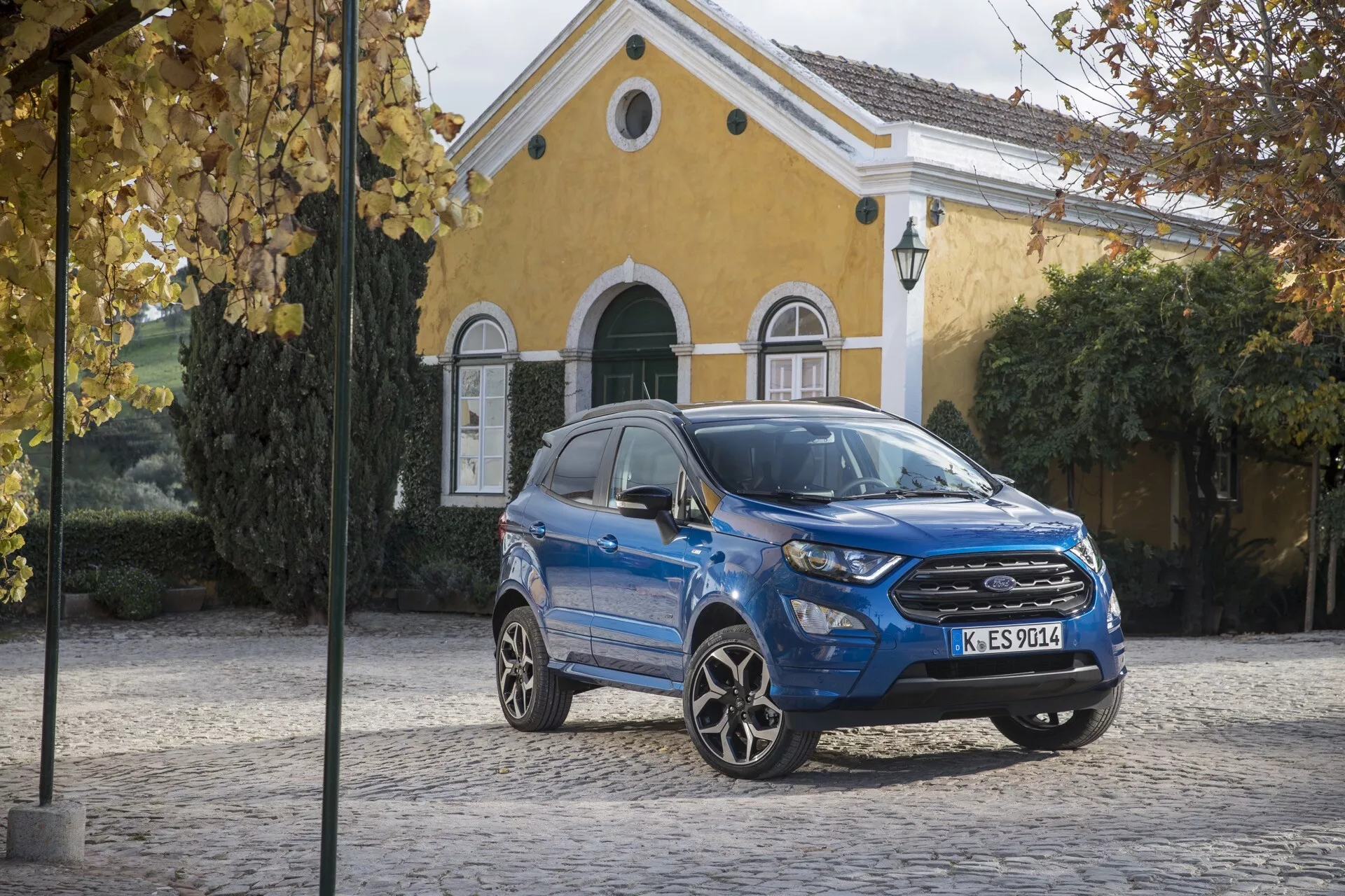 Niebieski kompaktowy SUV Ford EcoSport Facelift 2017 zaparkowany na bruku z żółtym budynkiem w tle, prezentujący przedni grill, LEDowe reflektory i sportowe felgi aluminiowe