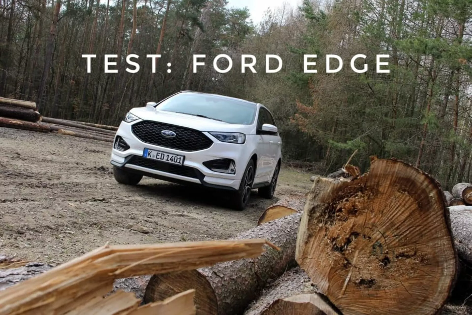 Biały Ford Edge 2018 pokazujący swoje zdolności terenowe z perspektywy trzech czwartych przedniej i bocznej, podkreślający LEDowe światła do jazdy dziennej, wieloramienne koła ze stopu i dwukolorowe malowanie, umieszczony na tle naturalnym z pociętymi kłodami drewna na pierwszym planie