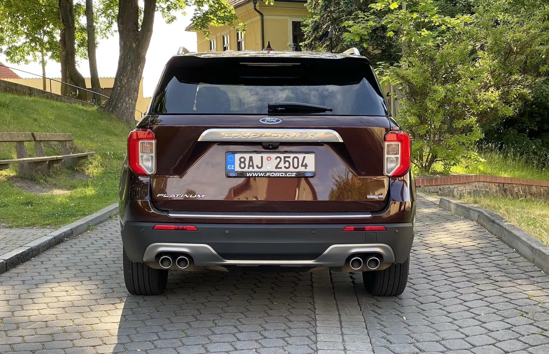 Tylny widok na SUV Ford Explorer 2019. Pojazd prezentuje elegancki tylny design z tylnymi światłami po obu stronach, emblematem Ford umieszczonym poniżej tylnej szyby, wysokim trzecim światłem stop i podwójnymi chromowanymi końcówkami wydechów. Dodatkowe cechy obejmują centralnie umieszczoną tablicę rejestracyjną z chromowanym paskiem powyżej, tylne czujniki w zderzaku sugerujące system wspomagania parkowania i możliwy bezdotykowy system otwierania bagażnika, wszystko uchwycone pod lekko niskim kątem.