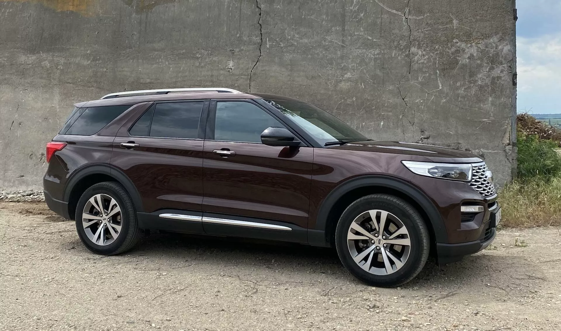 SUV Ford Explorer z 2019 roku zaparkowany na szorstkim żwirze w pobliżu betonowej konstrukcji, kąt z profilu bocznego podkreślający elementy designu takie jak duże felgi aluminiowe, wysoka linia boczna, przyciemniane szyby, klamki w kolorze nadwozia, chromowany pasek przy oknach, eleganckie linie nadwozia, lusterka z wbudowanymi kierunkowskazami, głęboki metaliczny lakier i zauważalny prześwit.