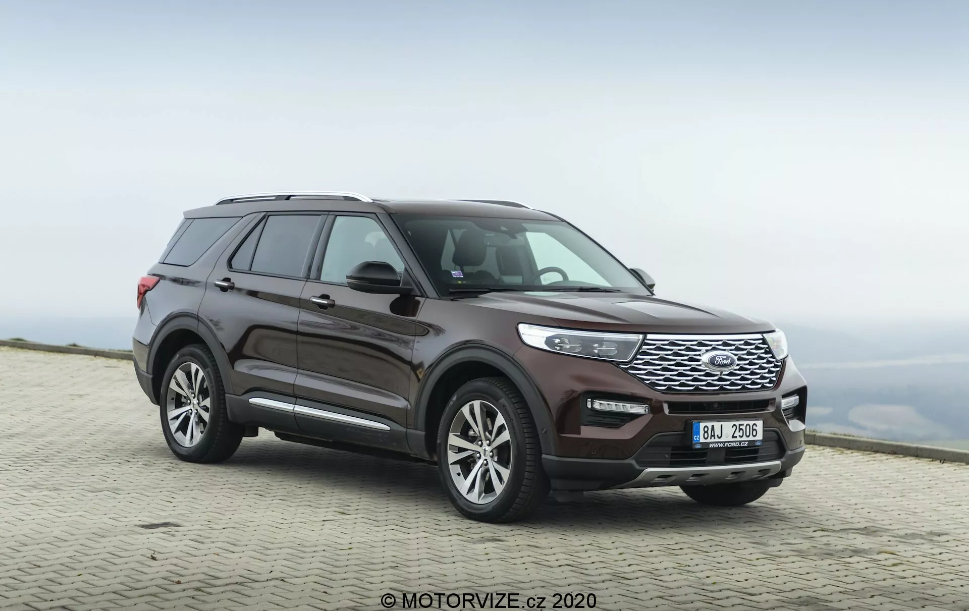 Zdjęcie SUV-a Ford Explorer 2019 uchwycone z przedniego trójczwartego kąta widzenia, podkreślając zarówno profil boczny, jak i przednią część pojazdu. Niski kąt fotografowania podkreśla solidną postawę i wyrazistą przednią kratę pojazdu. Widoczna jest cała długość samochodu, pokazując linie designu, wzór kół oraz kontury drzwi i błotników. Koła są lekko skręcone w prawo, co daje wrażenie ruchu. Wyróżniające się cechy to przednia krata z poziomymi listewkami, eleganckie reflektory zintegrowane z kratą oraz przedni zderzak z widocznymi czujnikami. Lusterka boczne wydają się mieścić zintegrowane kierunkowskazy, a linia dachu opada nieco ku tyłowi, z widocznymi relingami dachowymi. Wykończenie lakieru jest głębokie metaliczne w kolorze burgunda. Pojazd jest zaparkowany na utwardzonej powierzchni z rozmytym naturalnym tłem, co sugeruje dużą wysokość i pochmurne niebo.
