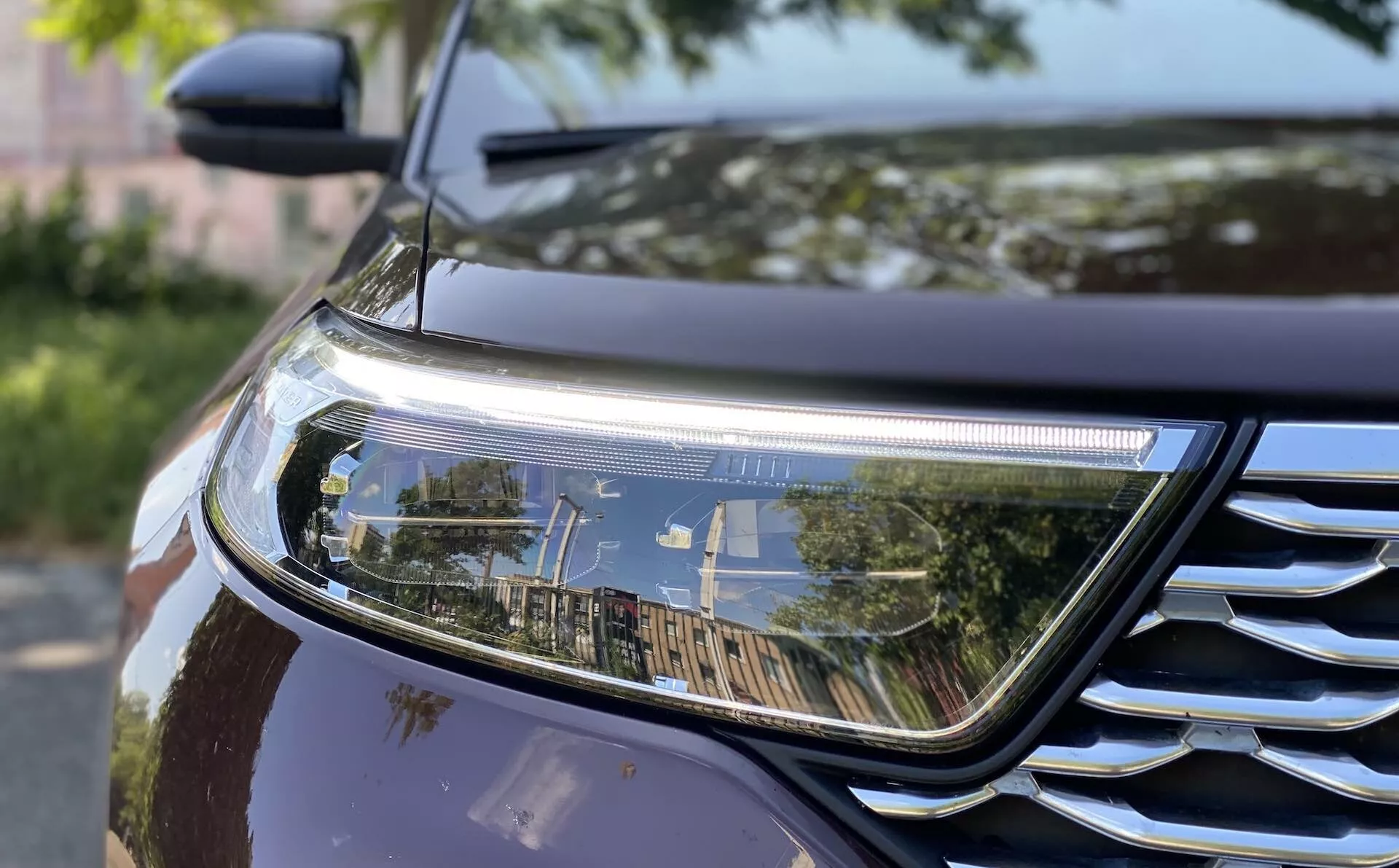 Zbliżenie przedniego fragmentu Forda Explorera 2019, pokazujące prawy reflektor i część kraty. Reflektor ma nowoczesny design z elementami LED lub HID i poziomym światłem dziennym, natomiast kratka ma chromowane akcenty z poziomymi listwami. Ciemna, metaliczna farba samochodu odbija otoczenie, podkreślając jego naturę na świeżym powietrzu. Obraz uwzględnia również część błotnika i maski, podkreślając aerodynamiczne kontury.