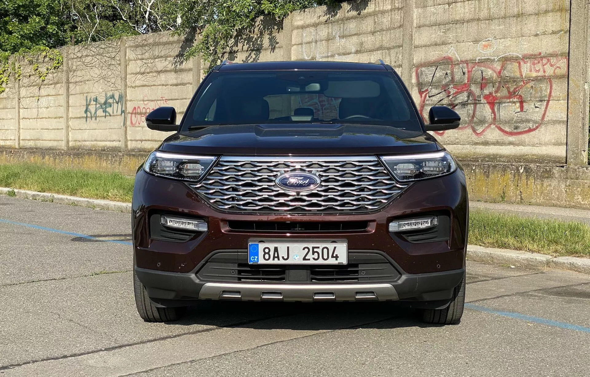Widok czołowy SUV-a Ford Explorer 2019, uchwycony pod kątem poziomym, prezentujący nowoczesny design pojazdu. Wyrazisty przedni panel charakteryzuje się dużą, chromowaną kratką w heksagonalny wzór i centralnie umieszczonym emblematem producenta. Smukłe, poziomo wydłużone reflektory LED oraz zderzak przedni w kolorze nadwozia z dolnymi wlotami powietrza i czujnikami parkowania podkreślają postęp technologiczny. Maskę zdobi kształtowany design z podwyższonymi liniami, podczas gdy błotniki i boczne lusterka z wbudowanymi kierunkowskazami dodają masywnego wyglądu. SUV, pomalowany na bogaty kasztanowy lub ciemnoczerwony kolor, zaparkowany jest na utwardzonym terenie z betonowym krawężnikiem i ścianą pokrytą graffiti w tle, podkreślając miejskie otoczenie.