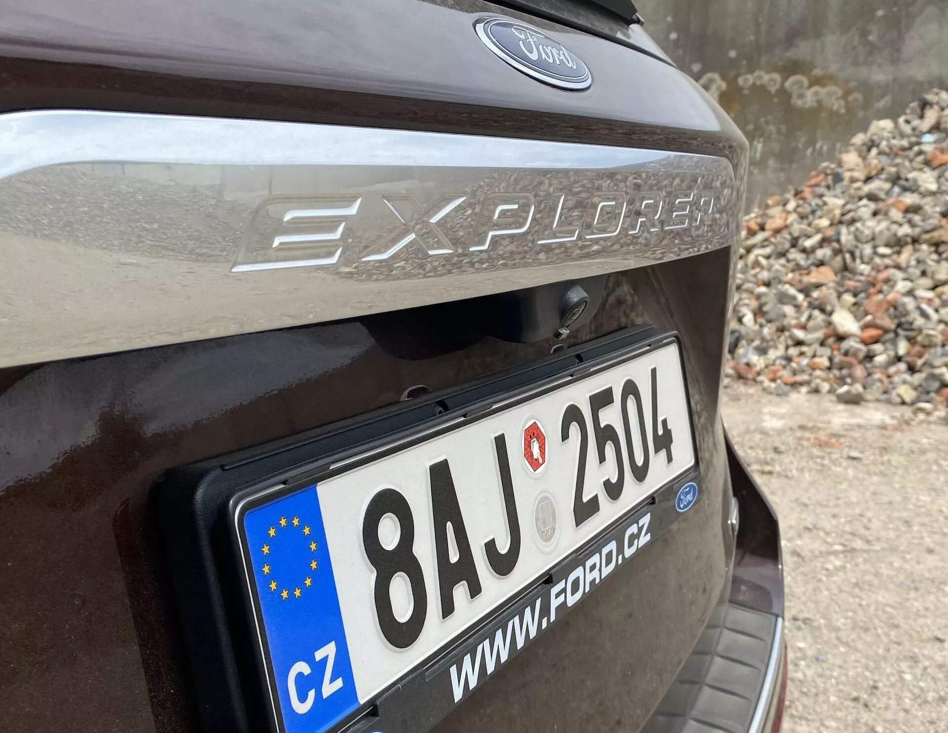 Zbliżenie na tylną część Forda Explorera z 2019 roku, skupiające się na obszarze tylnej klapy z wyraźnie widoczną tablicą rejestracyjną Unii Europejskiej z numerem '8AJ 2504' oraz kodem kraju 'CZ'. Nazwa modelu 'Explorer' i logo Forda znajdują się nad tablicą, a nad tablicą rejestracyjną widoczny jest pasek odblaskowy. Auto wydaje się być ciemnobrązowe lub ciemnoszare, zaparkowane w miejscu z kamieniami lub gruzem.