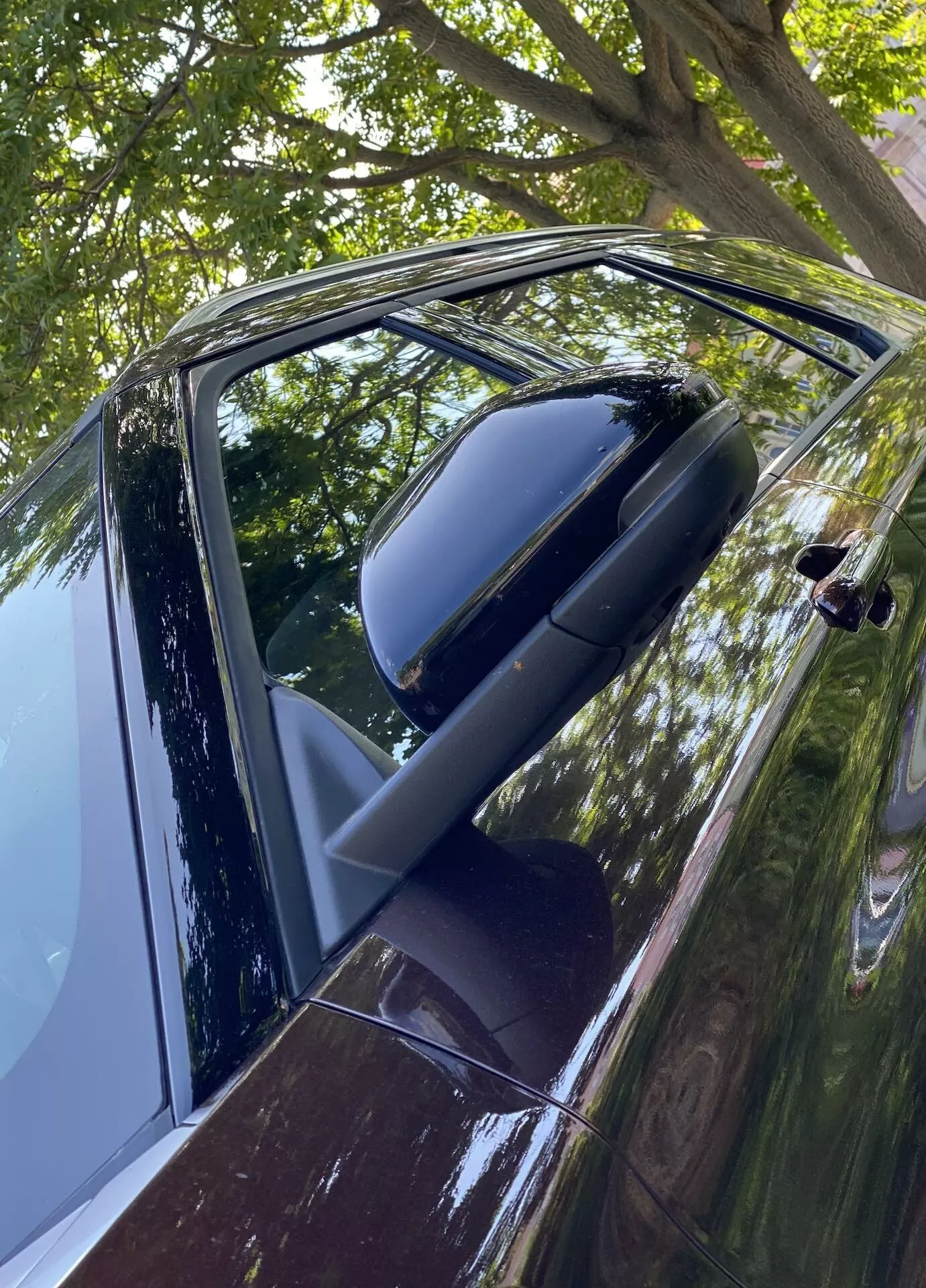Zbliżony, kątowny widok na drzwi kierowcy i lusterko boczne w Ford Explorer 2019. Zdjęcie wykonane nieco powyżej poziomu klamki drzwi odsłania błyszczący ciemny zewnętrzny wygląd, prawdopodobnie czarny lub głęboki niebieski, z odblaskami wskazującymi na czysty i zadbany nadwozie. Składane lusterko w kolorze nadwozia sugeruje model wyższej klasy. Częściowo opuszczona szyba i nowoczesna klamka, prawdopodobnie z czujnikiem dotykowym, sugerują technologię bezkluczykowego dostępu. Odbicia liści i gałęzi wskazują na parkowanie na zewnątrz.