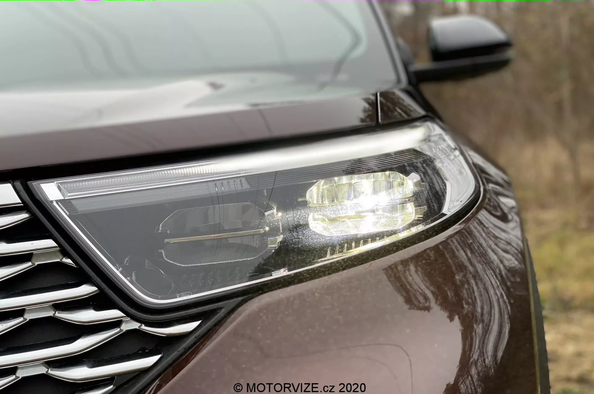Bliski widok boczny przedniego końca Forda Explorera 2019. Wyrazisty nowoczesny reflektor sugerujący technologię LED lub HID z aktywnymi światłami do jazdy dziennej. Częściowo widoczna kratka z metalicznym znaczkiem, umieszczona pod niskim kątem. Samochód ma ciemne, odbijające światło wykończenie lakieru, co wskazuje na metaliczny lub perłowy połysk, w warunkach zewnętrznych.