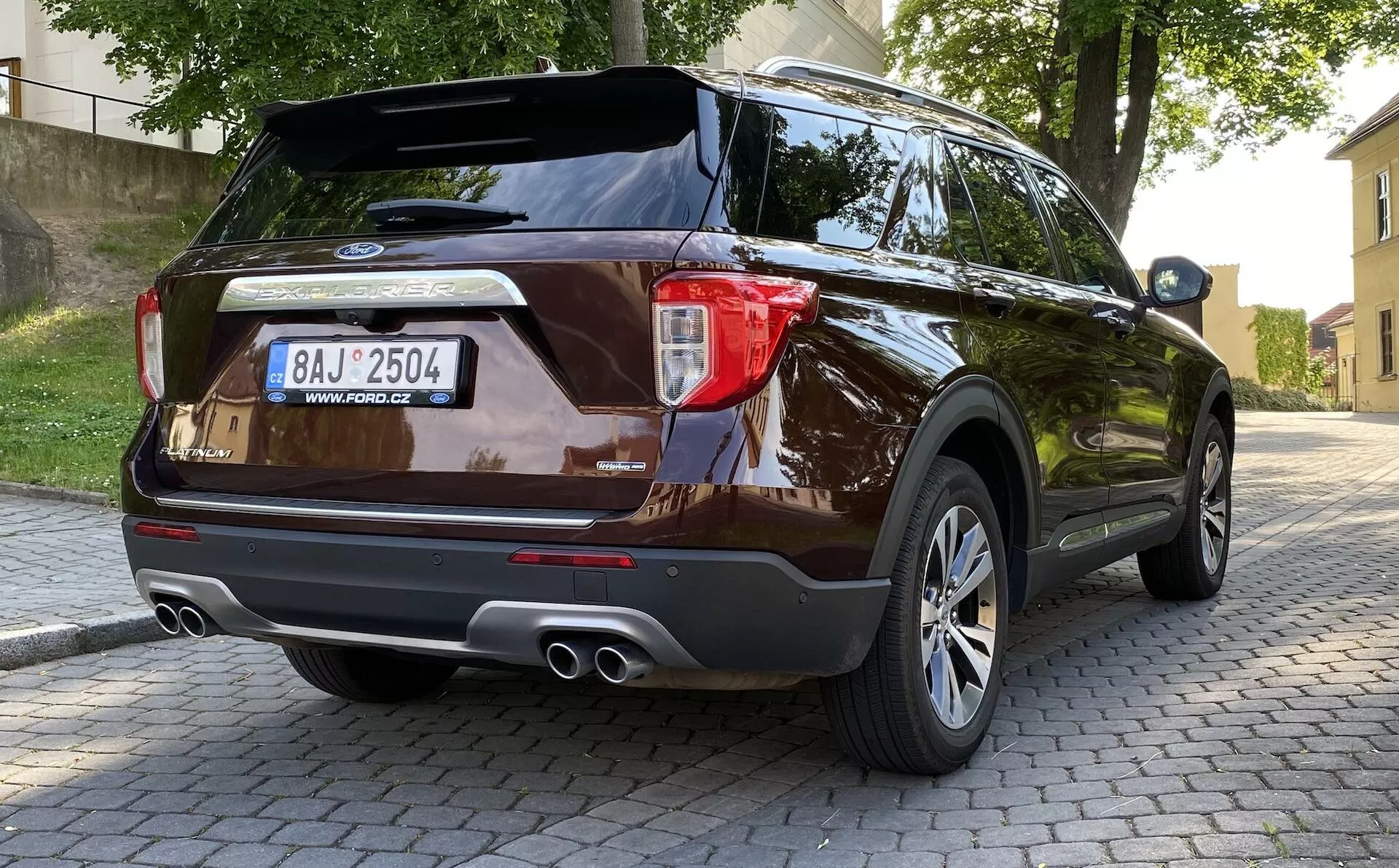 2019 Ford Explorer nowoczesne SUV z tylnej trzy-czwartej perspektywy pokazujące design tylnych świateł, tylny zderzak z czujnikami parkowania, podwójne rury wydechowe, wieloramienne felgi aluminiowe i ciemne metaliczne wykończenie lakieru, zaparkowane na brukowanej powierzchni w miejskim otoczeniu z roślinnością i budynkami w tle.