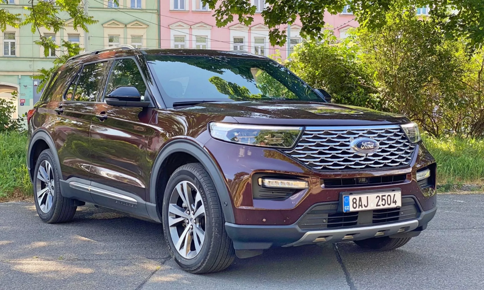 Widok z przodu i pod kątem na brązowego SUV-ę Ford Explorer 2019, ukazujący jego boczny profil i przednią fasadę. Samochód zaparkowany jest na miejskiej drodze, otoczonej budynkami i drzewami. Kluczowe widoczne cechy to wyrazista przednia krata z poziomymi listwami i centralnym emblematem, nowoczesne światła do jazdy dziennej LED, światła przeciwmgielne zintegrowane w dolnym zderzaku, duże wieloramienne alufelgi, lusterka boczne z wbudowanymi kierunkowskazami, przyciemniane szyby, relingi dachowe, profilowane linie nadwozia i podniesiony prześwit.