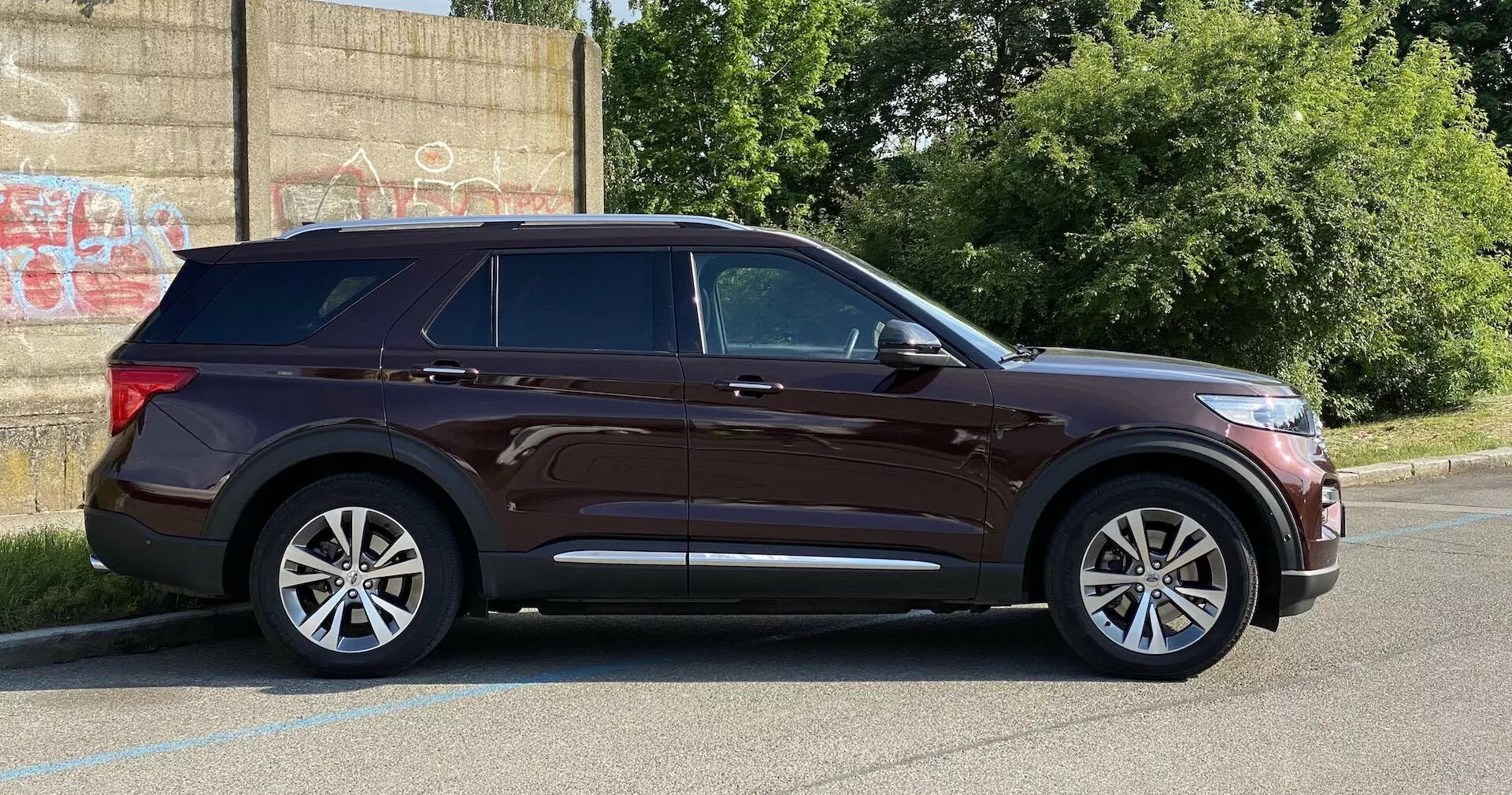 Nowoczesny SUV Ford Explorer 2019 widziany z boku, zaparkowany na utwardzonym placu obok krawężnika z graffiti na ścianie w tle, sugerującym miejskie otoczenie. Właściwości obejmują eleganckie reflektory, wyraźną linię charakterystyczną, lusterka boczne z kierunkowskazami, pięcioramienne alufelgi, chromowane detale wokół okien, relingi dachowe, przyciemniane tylne szyby, klamki w kolorze nadwozia i dużą odległość od podłoża. Fotografowanie w świetle dziennym pokazuje cały profil pojazdu.