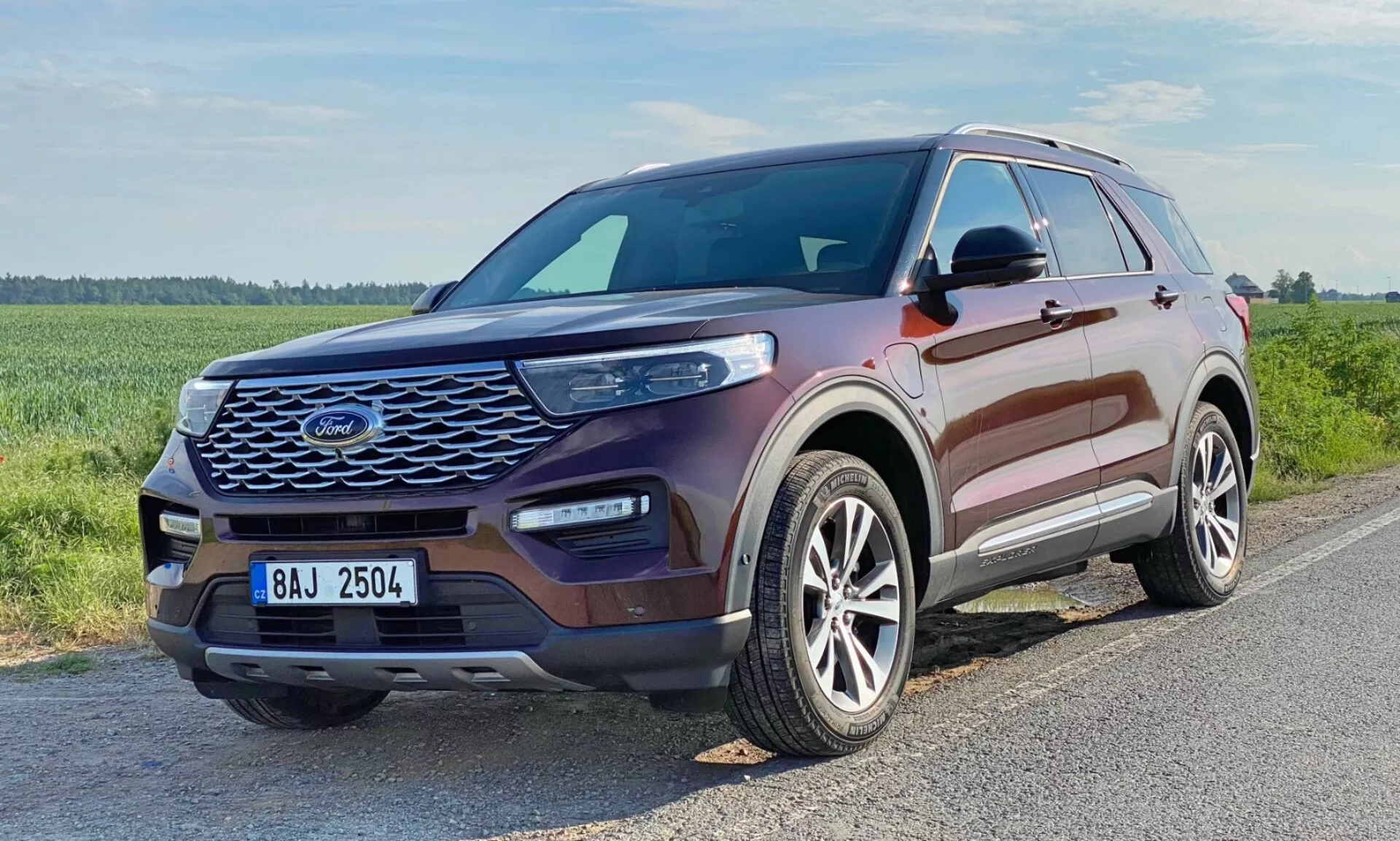 Trzy czwarte widoku z przodu samochodu SUV Ford Explorer 2019. Obraz przedstawia przedni grill pojazdu z wzorem siatki, smukłe wydłużone reflektory z widocznymi dziennymi światłami LED, rzeźbioną maską z podniesionymi konturami, aluminiowe koła z wieloramiennym wzorem oraz boczne lusterka dopasowane kolorystycznie do nadwozia z wbudowanymi kierunkowskazami. Okna mają chromowane lub srebrne obramowanie. Pojazd ma metaliczny, bordowy kolor i jest ustawiony na asfaltowej drodze w zielonej, potencjalnie wiejskiej okolicy.