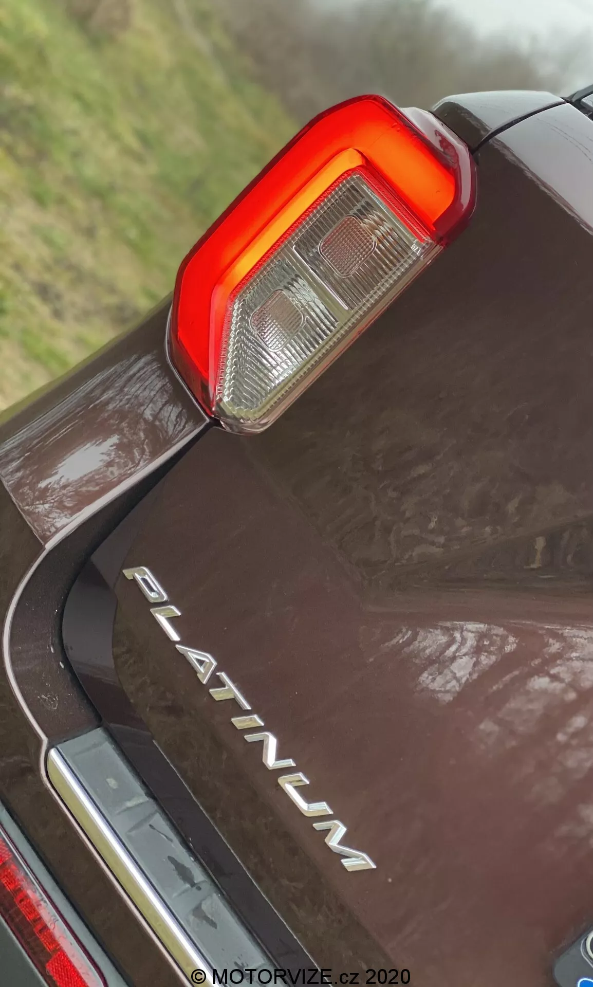 Zbliżenie, niski kąt tylnej części Forda Explorer 2019 w wersji PLATINUM, z naciskiem na pionowe tylne światło LED z przejrzystymi i czerwonymi sekcjami świateł cofania i hamowania, chromowaną plakietką 'PLATINUM', odbijającą ciemną farbą z kroplami wody i chromowanym elementem pod tylnym zderzakiem.