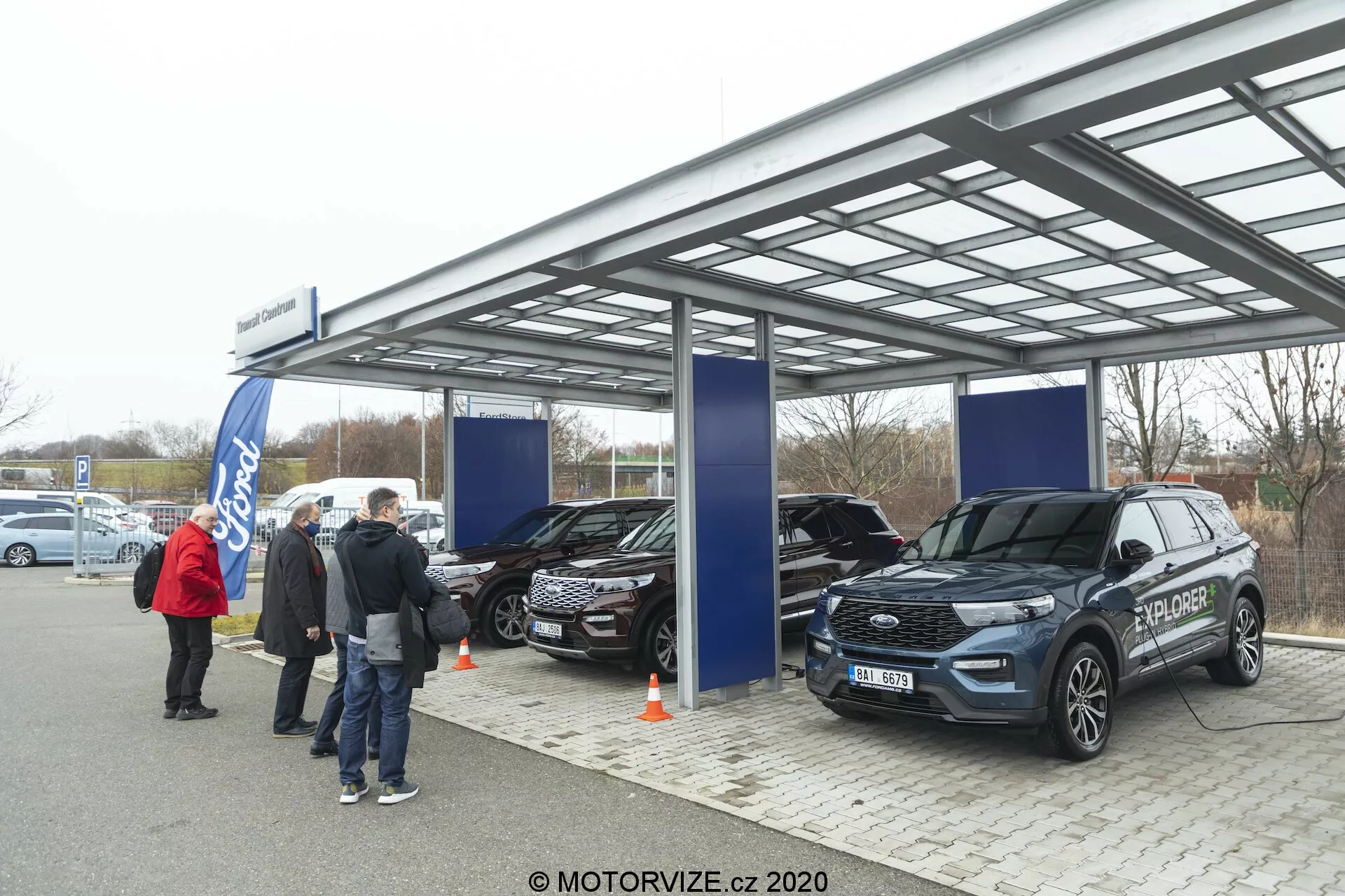 Obraz przedstawia zewnętrzną ekspozycję samochodową lub wydarzenie pod metalowym, półprzezroczystym dachem z miękkim, pochmurnym dziennym światłem. Trzy SUV-y, w tym nowoczesny Ford Explorer 2019 na pierwszym planie, są na pokazie z widocznymi cechami jak przedni grill, reflektory i lusterka boczne. Ludzie i bannery sugerują oficjalną imprezę promocyjną, a logo Forda jest widoczne w tle.
