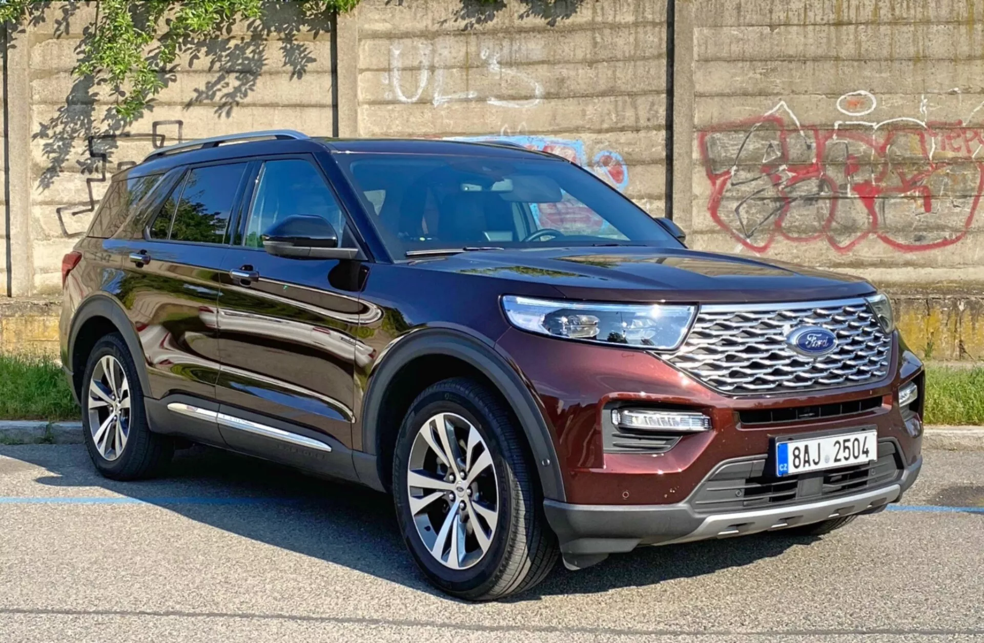Zdjęcie SUV Ford Explorer 2019 wykonane pod kątem trzech czwartych z przodu od strony kierowcy, pokazujące błyszczący metaliczny brązowy lub burgundowy kolor. Zdjęcie podkreśla przedni grill z logo Ford, eleganckie światła LED lub HID, przedni zderzak i profil boczny kierowcy, w tym drzwi, błotnik i wieloramienne felgi aluminiowe. Pojazd jest zaparkowany w miejskiej okolicy z graffiti na betonowej ścianie w tle, podkreślając jego solidny i nowoczesny design, z możliwymi zaawansowanymi systemami wspomagania kierowcy, takimi jak adaptacyjny tempomat i asystent utrzymania pasa ruchu.