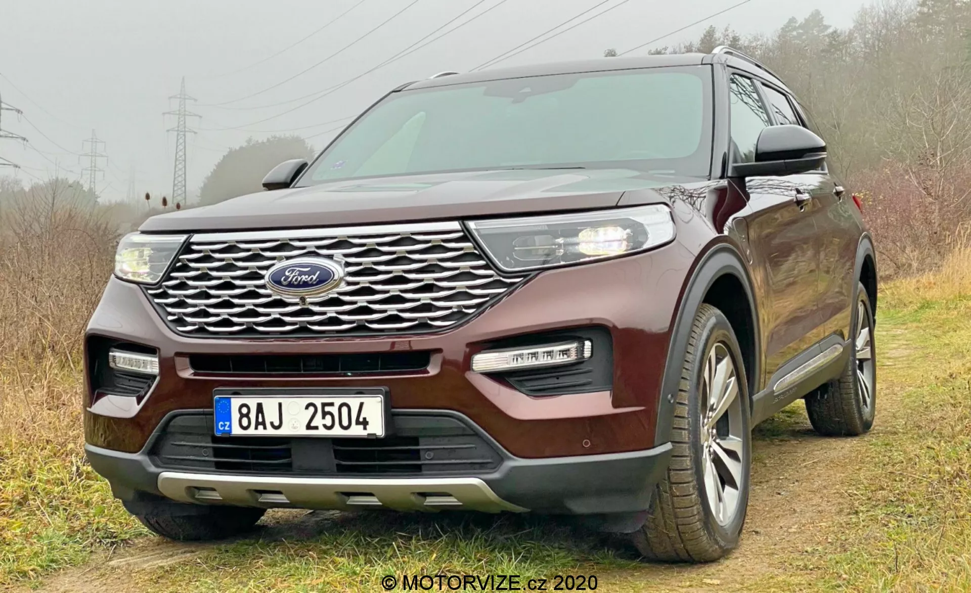 Ford Explorer 2019 samochód sfotografowany pod niskim przednim kątem trzy czwarte, ukazując kratkę z logo Forda, reflektory, światła przeciwmgielne, maskę, boczne szyby i część tylnej części. Wysublimowany design kratki, eleganckie reflektory LED, dwukolorowy zderzak i bezkluczykowy system wejścia podkreślają nowoczesność i zaawansowane technologie. Ustawiony nieco na drodze terenowej z prawymi kołami na trawie i lewymi kołami na ziemi, podkreślając zdolności do lekkiej jazdy terenowej. Widoczne wieloszprychowe felgi aluminiowe, wskazujące na opony całoroczne.