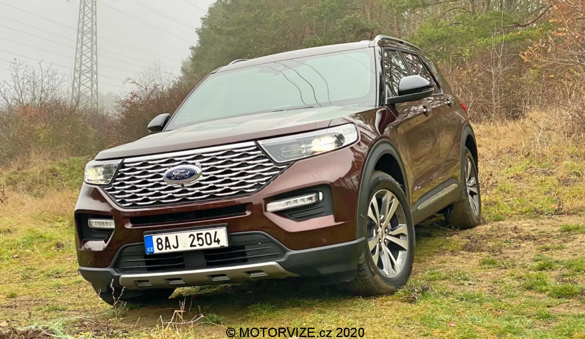 Przedni, trójkątny widok SUV-a Ford Explorer 2019 zrobiony pod niskim kątem, pokazujący jego wyraźny chromowany grill, eleganckie reflektory LED i wieloszprychowe felgi aluminiowe. SUV stoi na nieutwardzonej powierzchni z roślinnością, sugerując jego zdolność do jazdy terenowej. Przedni zderzak zawiera światła przeciwmgielne i wloty powietrza, sugerując ulepszoną funkcjonalność. Elementy takie jak czujniki lub kamery mogą wskazywać na zaawansowane funkcje bezpieczeństwa. Tablica rejestracyjna jest ocenzurowana, a w prawym dolnym rogu znajduje się znak wodny '© MOTORVIZE.cz 2020'. Kadr uwydatnia jego solidną postawę i zaawansowaną technologię.