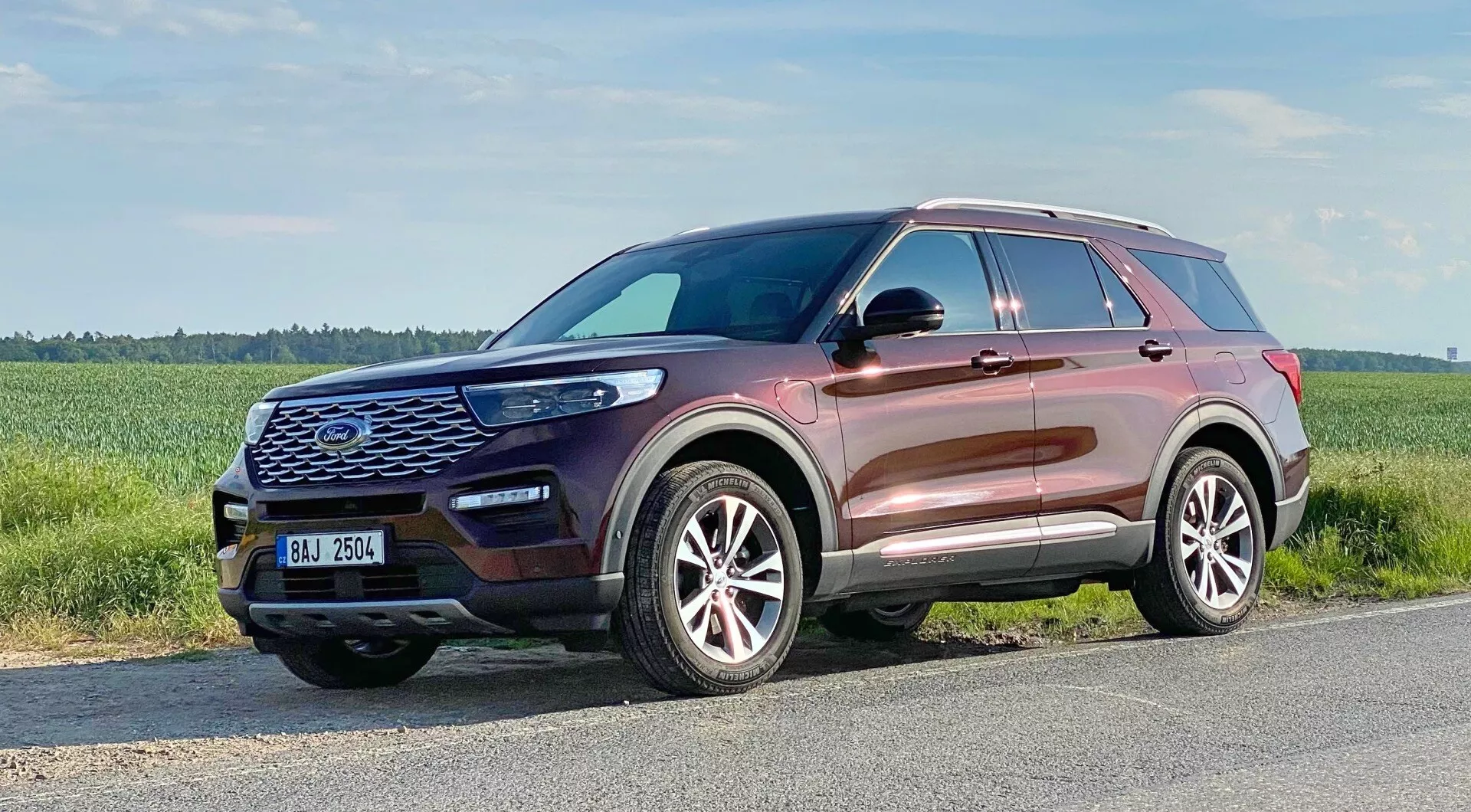 Ford Explorer 2019 - Nowoczesny SUV uchwycony z trzech czwartych frontalnego widoku, podkreślający przedni grill z sześciokątnym wzorem i dużym emblematem, konturowaną maskę, eleganckie reflektory LED, światła przeciwmgielne, wloty powietrza, wieloramienne aluminiowe felgi, lekko wznoszącą się linię okien, relingi dachowe, lusterka boczne z zintegrowanymi kierunkowskazami i metaliczny kolor nadwozia odbijający wysokiej jakości wykończenie.