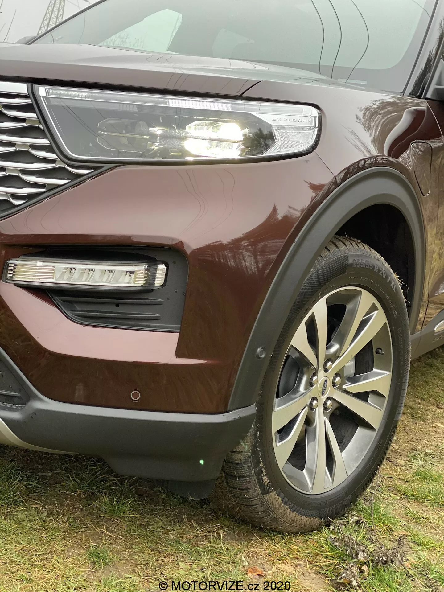 Zbliżenie z kąta na SUV Ford Explorer 2019, skupiające się na przedniej lewej ćwiartce, z akcentem na duże koła ze stopu metali lekkich, z grillem z chromowanymi akcentami oraz częściowo widocznym reflektorem LED ze światłami do jazdy dziennej, metaliczną burgundową farbą, światłem przeciwmgielnym w zagłębionej części zderzaka o innej teksturze i ewentualnym wlotem powietrza lub ozdobnym listwą. Logo znajduje się na środku kół wieloramiennych. Dolna część zderzaka sugeruje obecność czujnika parkowania lub systemu adaptacyjnego tempomatu. Zdjęcie wykonane z niskiego kąta, podkreślając wielkość i solidność pojazdu, z widocznym w prawym dolnym rogu znakiem wodnym 'MOTORVIZE.cz 2020'.