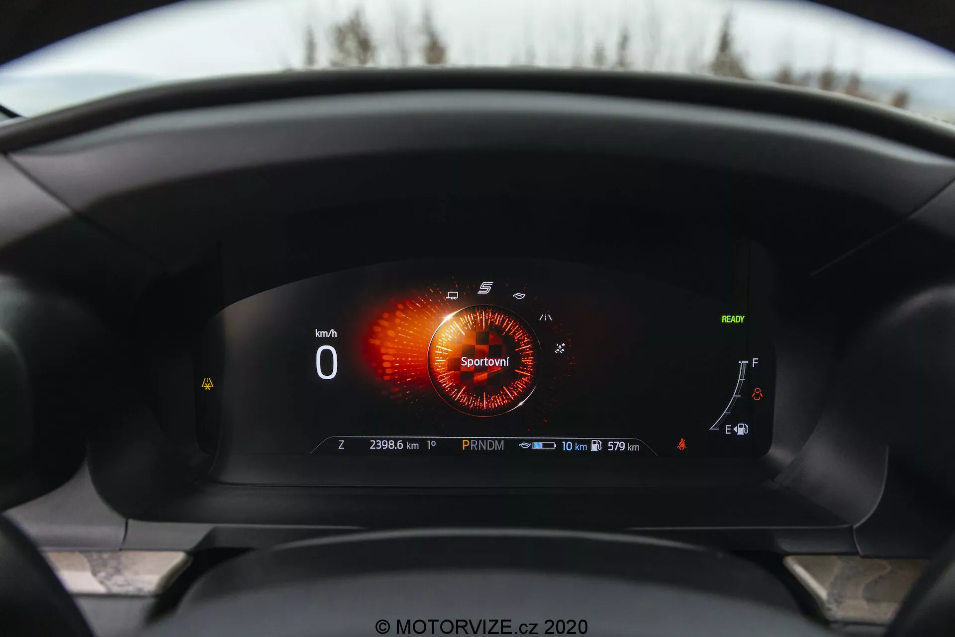 Widok wnętrza Forda Explorer 2019 z perspektywy kierowcy, prezentujący w pełni cyfrowy panel instrumentów. Wyświetlacz cyfrowy pokazuje obrotomierz w trybie 'Sport' z czerwono-pomarańczową grafiką, prędkościomierz pokazujący '0 km/h,' prawie pełny wskaźnik paliwa, wskaźnik temperatury chłodziwa na dolnym końcu i różne wskaźniki. Można zauważyć symbol adaptacyjnego tempomatu, przypomnienie o interwale serwisowym mówiące '10 000 km lub 365 dni,' oraz licznik przebiegu na '23063 km,' co wskazuje, że samochód stoi w trybie 'Park.' Prawie pełna ikona baterii sugeruje zdolności hybrydowe lub elektryczne.