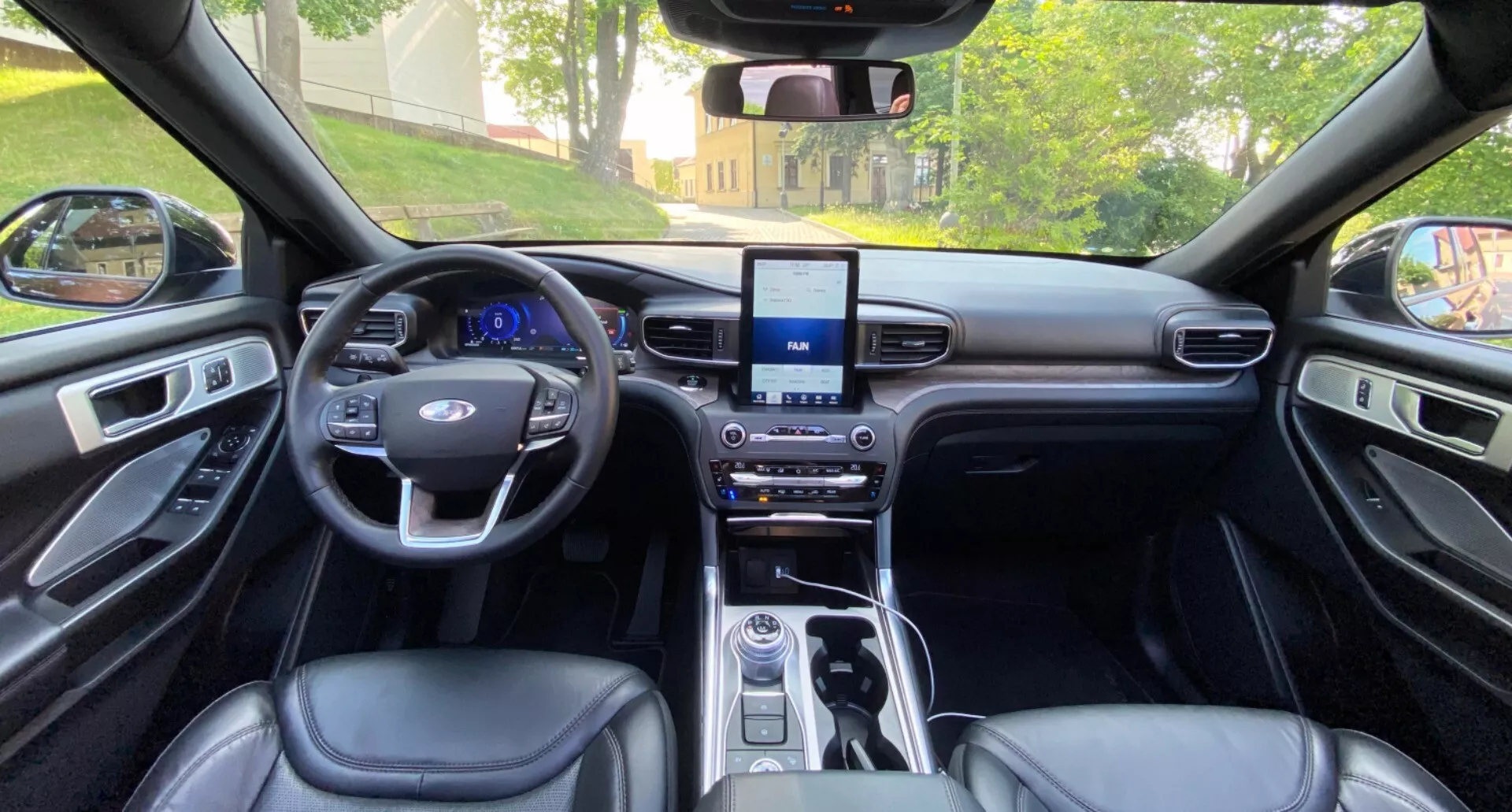 Wnętrze Forda Explorer 2019 widziane z perspektywy tylnych siedzeń, skierowane w kierunku deski rozdzielczej. Obraz przedstawia symetryczny układ, w tym kierownicę z przyciskami wielofunkcyjnymi, dotykowy ekran systemu infotainment, konsolę zmiany biegów ze sterownikami oraz tapicerkę siedzeń i paneli drzwi przypominającą skórę. Wnętrze premium jest podkreślone eleganckimi metalowymi wykończeniami i dobrze rozmieszczonym oświetleniem.