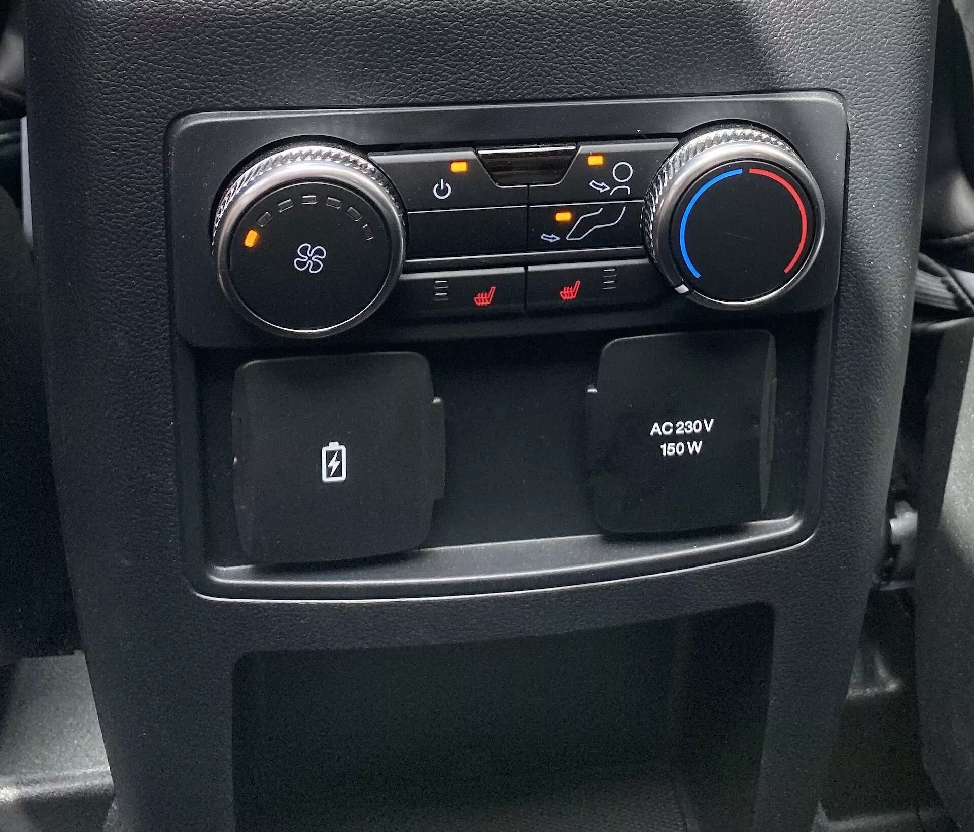 Zbliżenie sekcji konsoli środkowej wnętrza samochodu Ford Explorer 2019, prezentujące sterowanie HVAC i komfortu. Po lewej stronie znajduje się okrągłe czarne pokrętło z ikoną wentylatora do regulacji dmuchawy; obok niego przyciski do kierunku przepływu powietrza, recyrkulacji powietrza i funkcji odmrażania szyb. Po prawej stronie pokrętło w kolorach niebieskim i czerwonym do regulacji temperatury. Poniżej znajdują się dwa antypoślizgowe schowki, jeden zaprojektowany na kartę kluczową, oraz gniazdo zasilania AC po stronie pasażera oferujące 230V i 150W do urządzeń elektronicznych.
