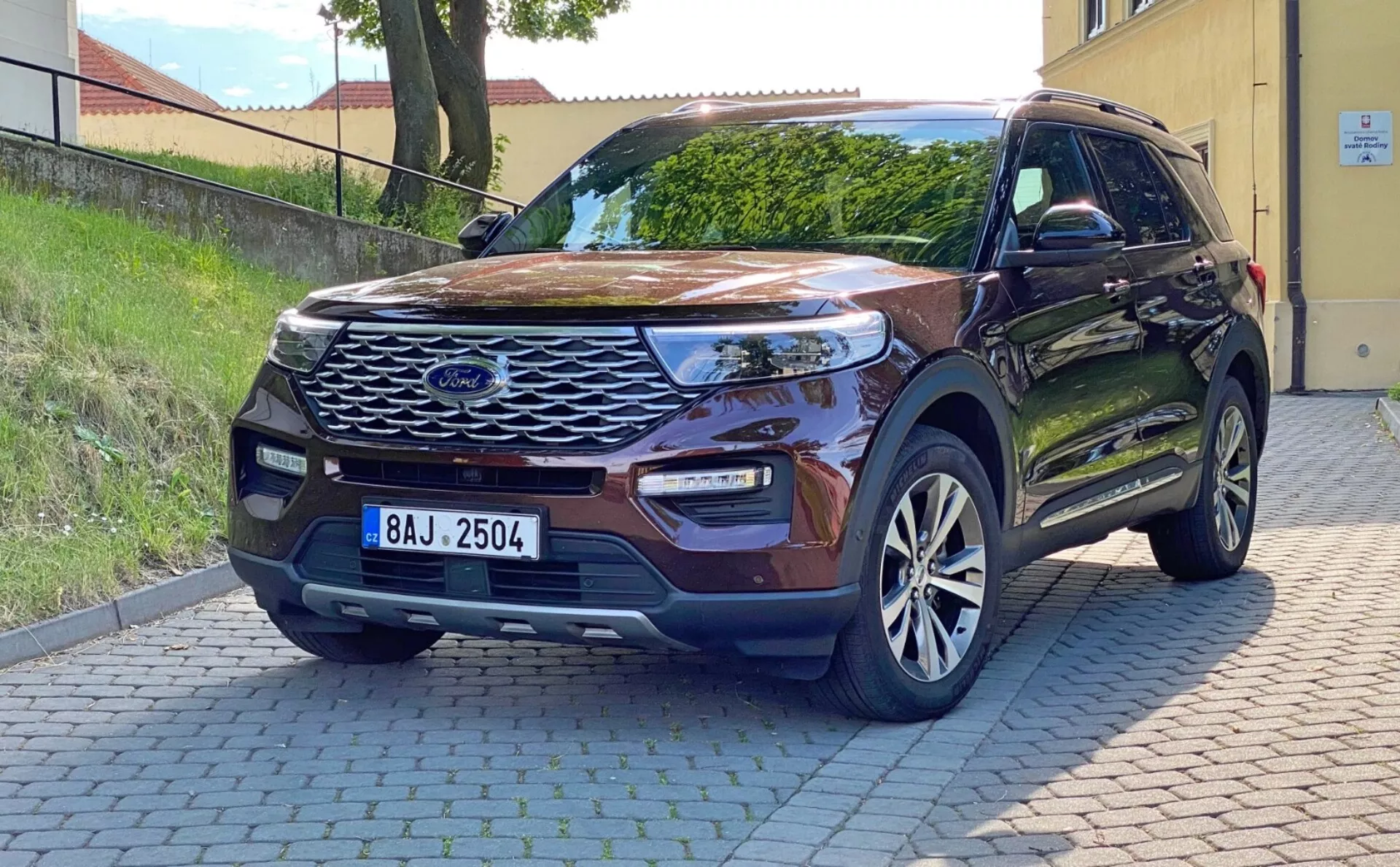 Zdjęcie nowoczesnego SUV-a Ford Explorer 2019 zaparkowanego na utwardzonej powierzchni, uchwycone z perspektywy przedniego trójtrzeciego ujęcia. Pokazuje wyraźny przedni grill z centralnym emblematem, smukłe przednie reflektory LED, zintegrowane światła przeciwmgielne w zderzaku, czujniki parkowania, rzeźbioną maskę, felgi aluminiowe, lusterka boczne w kolorze nadwozia z kierunkowskazami, bezkluczykowy dostęp i tło mieszkalne. Naturalne światło dzienne podkreśla cechy zewnętrzne i postęp technologiczny pojazdu.