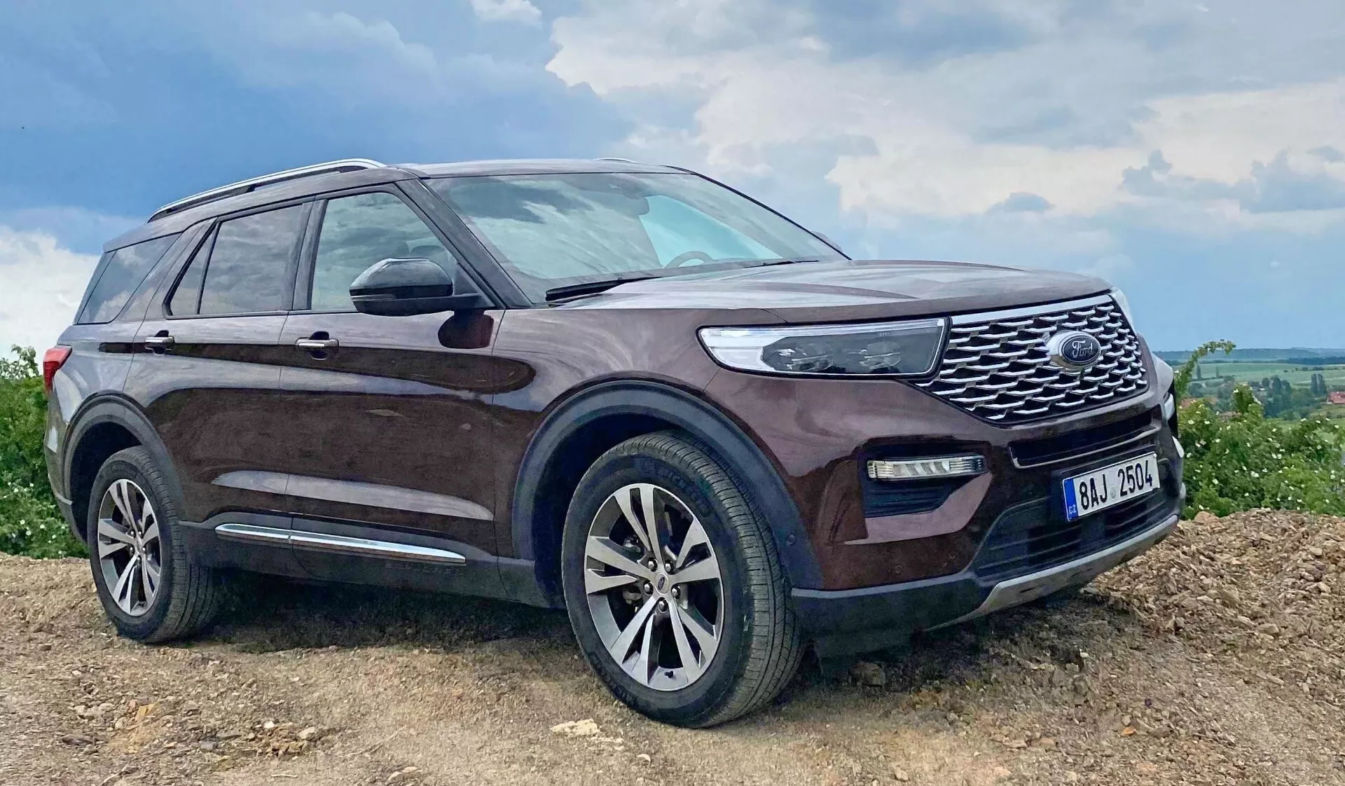 Obraz SUV-a Ford Explorer 2019 zaparkowanego na pochyłej, prawdopodobnie nieutwardzonej powierzchni, z ciemnym odcieniem i zabłoconymi bokami, wyrazistą przednią kratką z siatką, reflektorami LED, światłami przeciwmgielnymi, aluminiowymi felgami z wieloszprychowym wzorem, lusterkami bocznymi z zintegrowanymi kierunkowskazami, relingami dachowymi, bocznym stopniem, lekko przyciemnionymi szybami i anteną na tylnej części dachu, podkreślając jego wytrzymałe możliwości i luksusowe cechy w wiejskim lub off-road’owym otoczeniu.