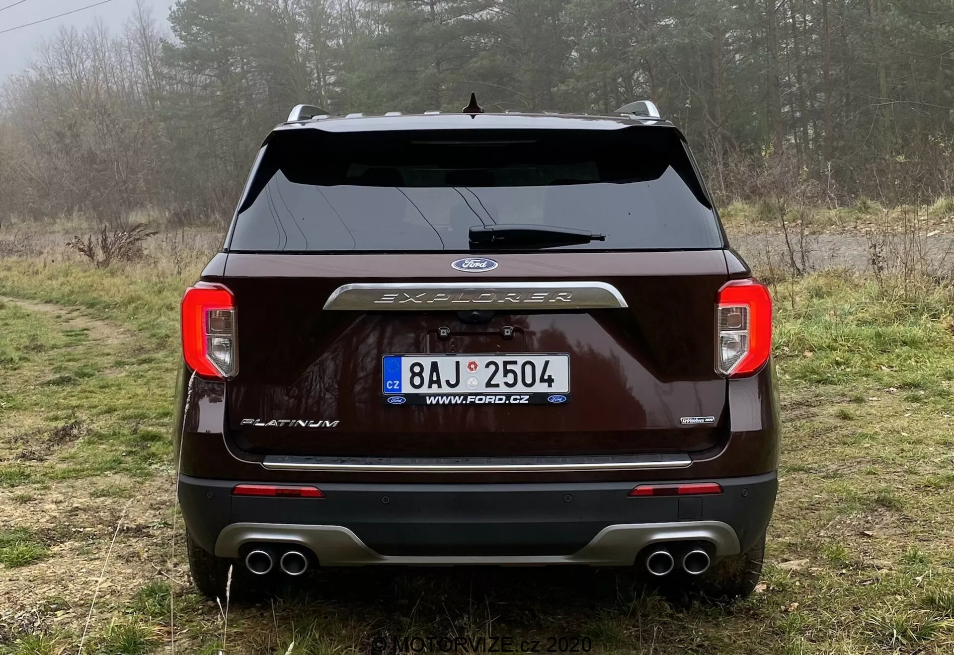 Tylny widok ciemnego Forda Explorera 2019 zaparkowanego na polnej drodze w wiejskim krajobrazie z roślinnością i mgłą. Obrazek pokazuje tylne światła z przejrzystymi soczewkami, centralnie umieszczone logo Ford, napis 'EXPLORER', wycieraczkę tylnej szyby, czujniki parkowania, system podwójnego wydechu i osłonę haka holowniczego. Widać tablicę rejestracyjną z czeską domeną, podkreślającą zewnętrzny design i technologię pojazdu.