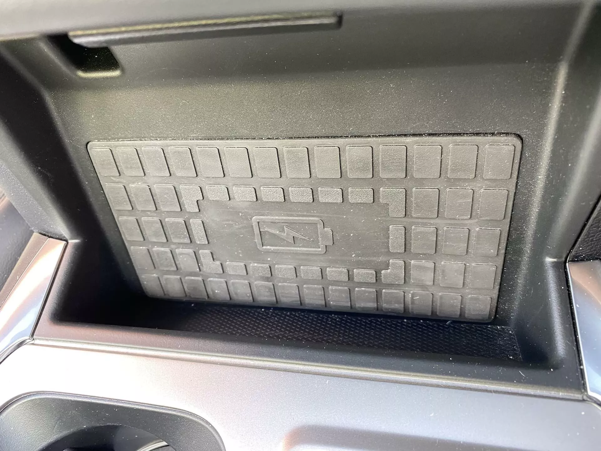 Zbliżenie na schowek konsoli centralnej Ford Explorer 2019, podkreślający bezprzewodową podkładkę ładującą o wzniesionej, kratkowanej fakturze i antypoślizgową powierzchnię. Widoczne są lśniące czarne plastikowe obramowania, dodające nowoczesnego stylu wnętrza samochodu. Światło słoneczne sugeruje zdjęcie wykonane w ciągu dnia z przedniego siedzenia, a symbol ładowania oznacza zaawansowaną technologię ładowania bezprzewodowego.