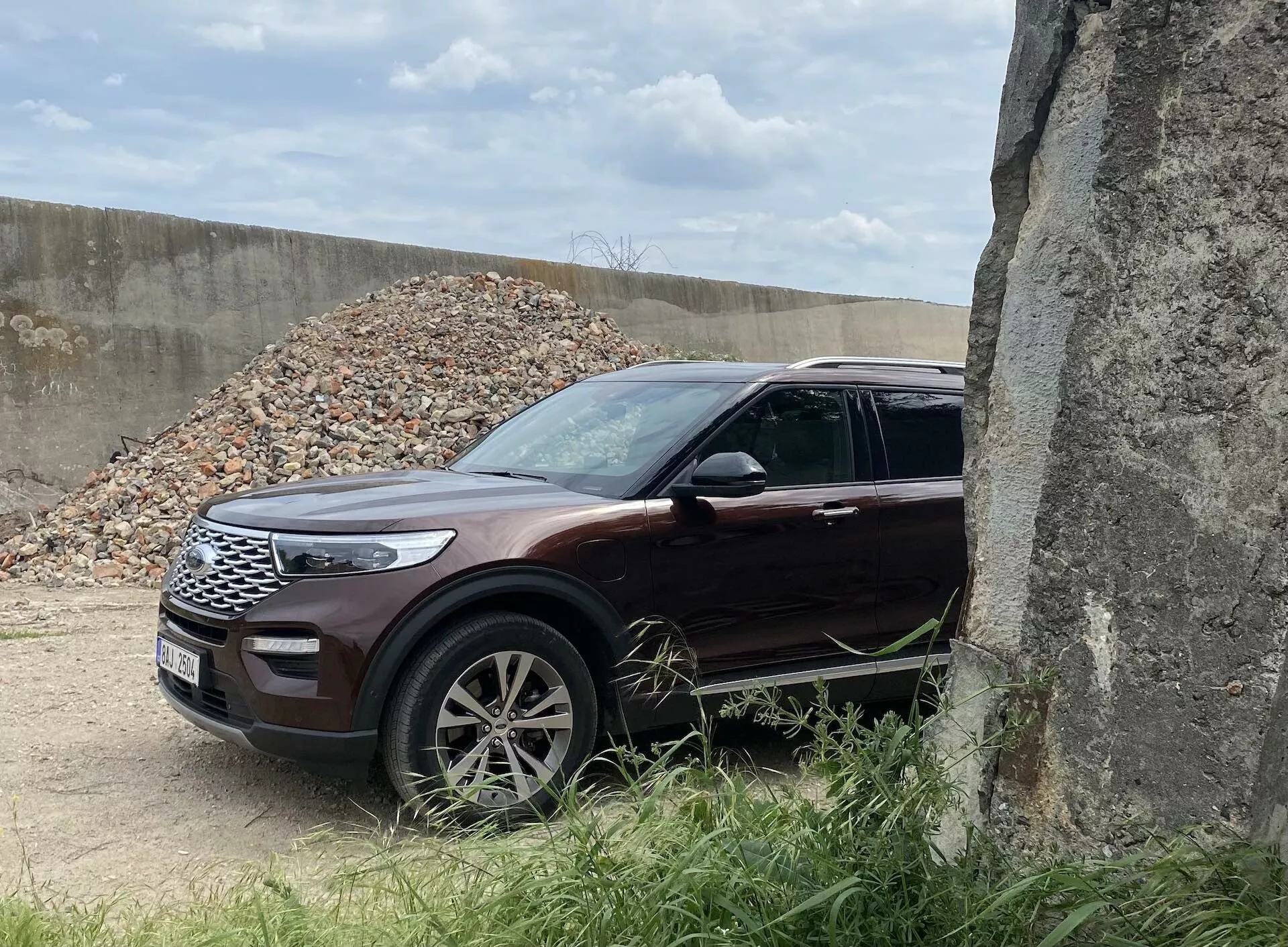 Obraz przedstawia SUV-a Ford Explorer z 2019 roku z trzech czwartych przedniego widoku, podkreślając przedni lewy grill, eleganckie reflektory LED, maskę, przednie koło i błotnik. Pojazd, w błyszczącym głębokim bordowym lub burgundowym kolorze, znajduje się w plenerze z żwirem w tle, sugerującym plac budowy. Wieloramienne felgi, zintegrowane kierunkowskazy w lusterkach i chromowane wykończenia dodają prestiżowego wyglądu. Częściowo zasłonięty betonową konstrukcją po prawej stronie, ten SUV charakteryzuje się nowoczesnym designem i elementami bezpieczeństwa, czyniąc go odpowiednim do kategorii takich jak SUV, nowoczesny design samochodowy, technologia pojazdów i pojazdy terenowe.