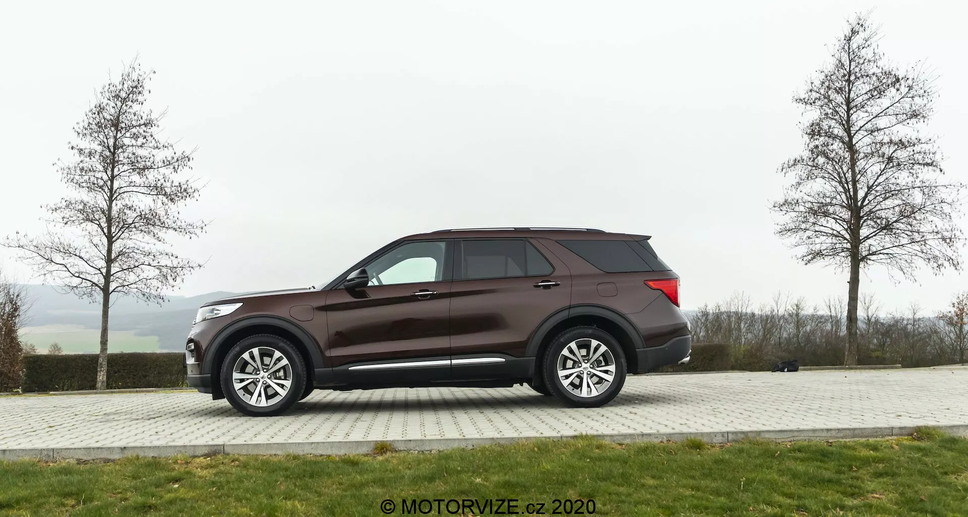 Widok boczny SUV-a Ford Explorer z 2019 roku zaparkowanego na utwardzonym terenie z trawą w tle w pochmurny dzień. Pojazd ma duże, wieloszprychowe felgi aluminiowe, przyciemniane szyby i ciemnobrązowy lub bordowy lakier. Elementy wyposażenia obejmują prawdopodobnie regulowane elektrycznie lusterka boczne z zintegrowanymi kierunkowskazami, aerodynamiczną linię dachu i widoczną antenę z tyłu.