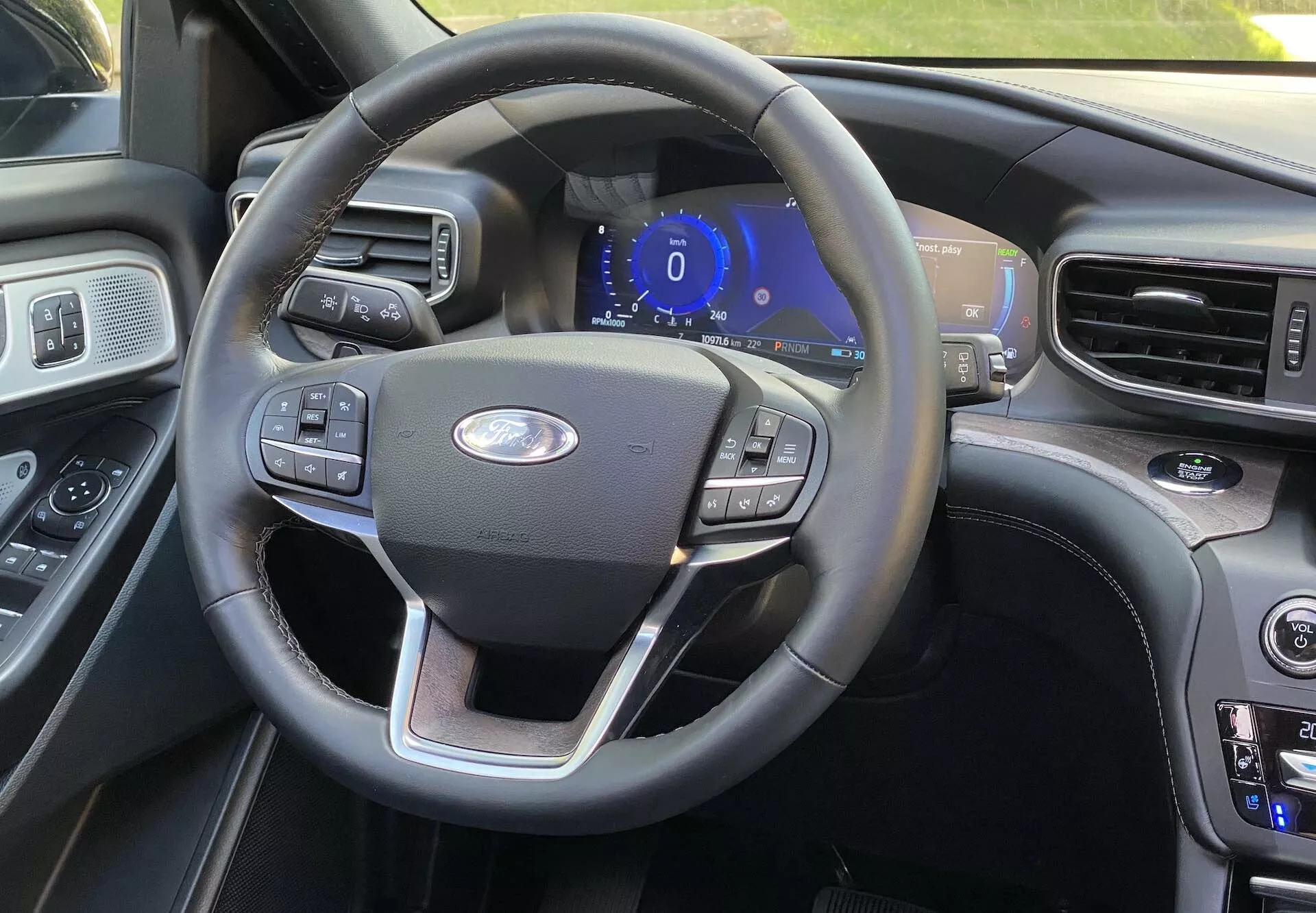 Widok wnętrza Ford Explorer 2019 z miejsca kierowcy, pokazujący kierownicę trzema ramionami z wielofunkcyjnymi przyciskami do tempomatu, ustawień audio i łączności telefonicznej. Centralnie umieszczone logo Forda oraz cyfrowy panel instrumentów podświetlony na niebiesko sugerują, że pojazd jest w trybie akcesoriów lub trybie startowym. Po prawej widać część konsoli środkowej z przyciskami do sterowania klimatyzacją i ustawień temperatury, co wskazuje na podwójną klimatyzację. Wnętrze zawiera połączenie skórzanych i drewnianych wykończeń, co tworzy luksusowe wrażenie, z kolejnym, większym ekranem lekko widocznym na krawędzi.