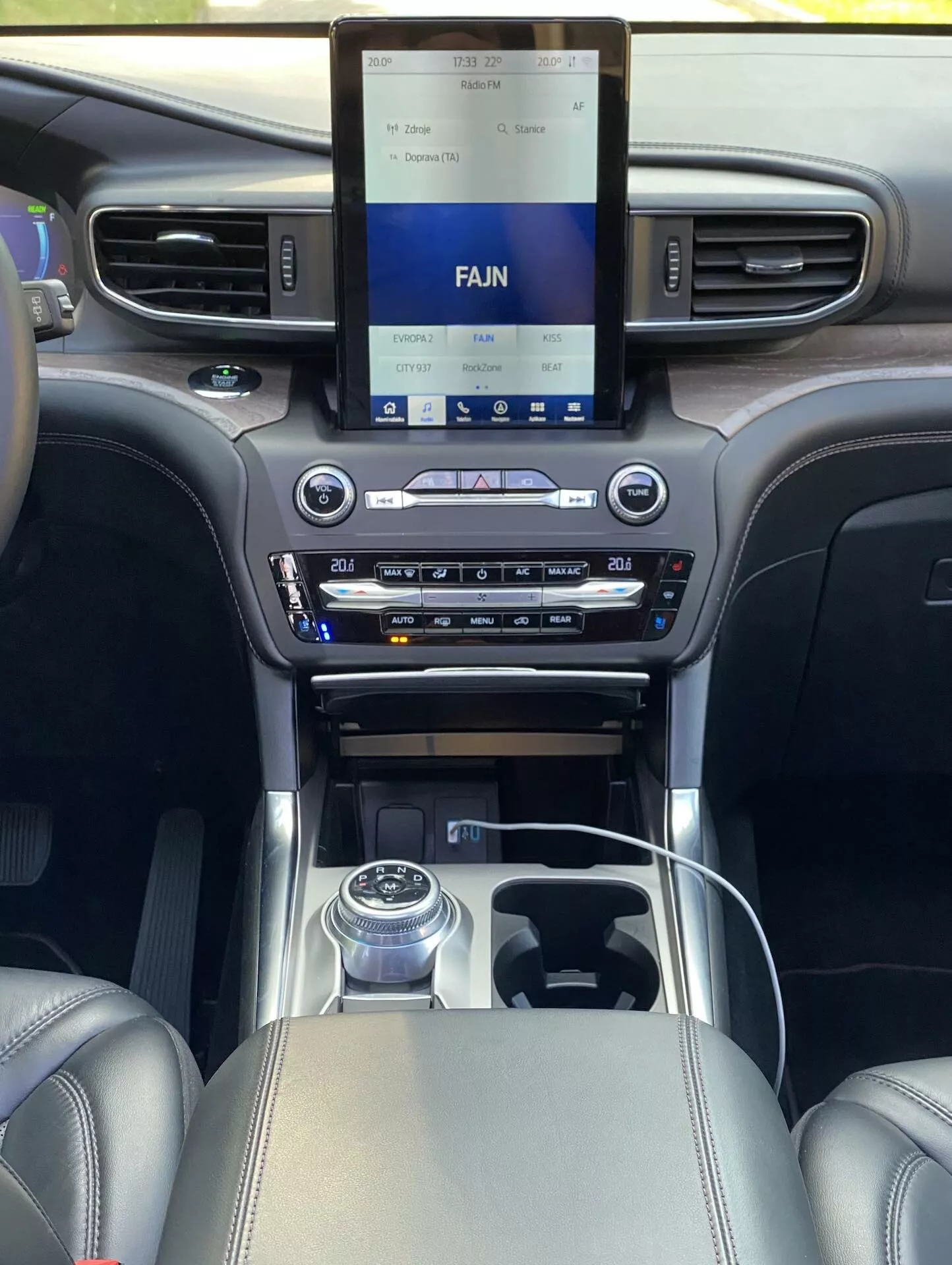 Widok wnętrza Forda Explorer 2019 z perspektywy pasażera siedzącego z tyłu, z naciskiem na przednią kabinę z zaawansowanym systemem informacyjno-rozrywkowym na konsoli centralnej, dwustrefową klimatyzacją, obrotowym pokrętłem zmiany biegów i luksusowymi detalami ze skóry i chromu.