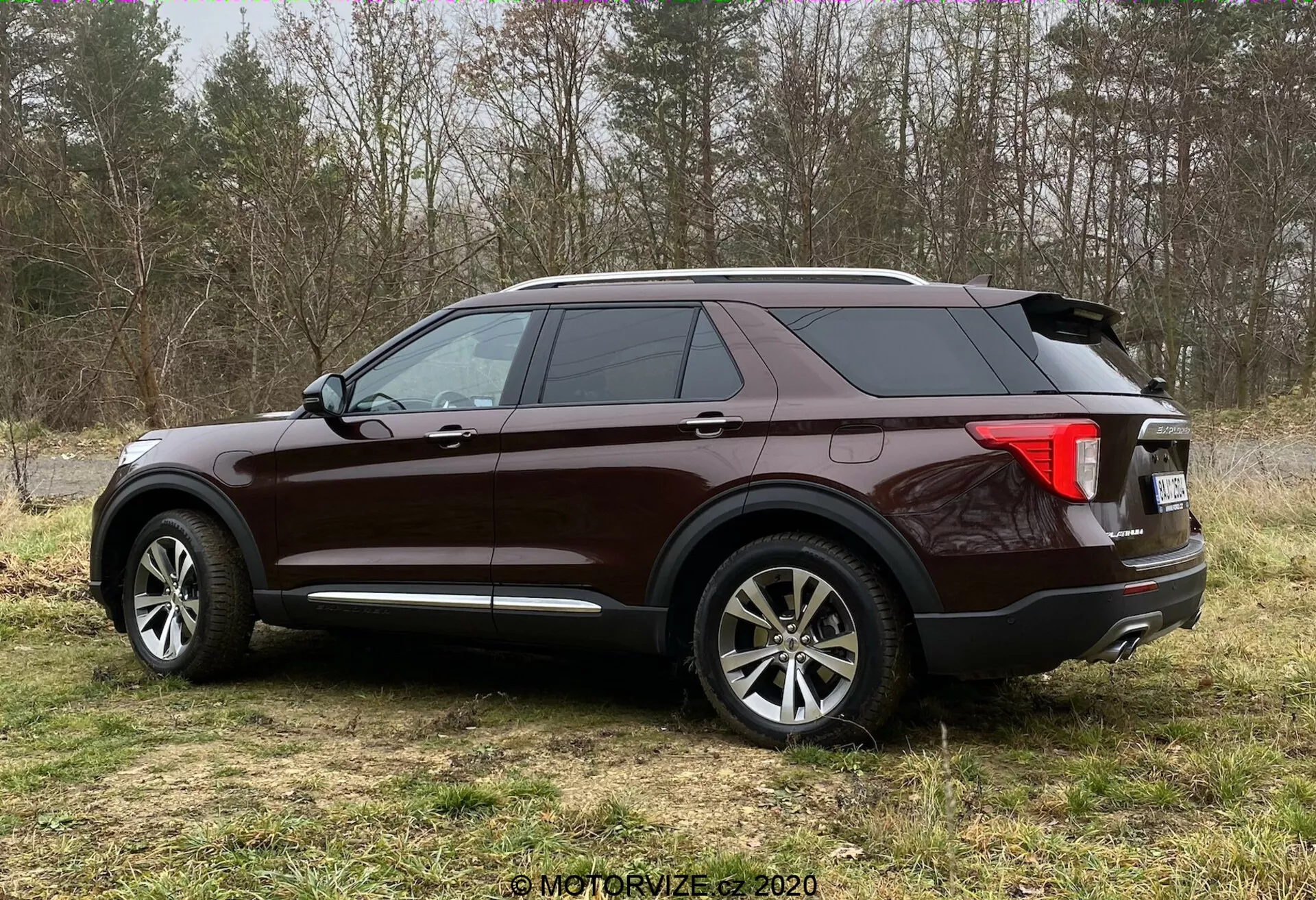 Widok boczny SUV-a Ford Explorer 2019 w błyszczącym burgundowym wykończeniu, ustawiony na trawiastej powierzchni z półleśnym tłem. Kąt trzy czwarte pokazuje duże, wieloramienne koła aluminiowe, opadającą linię dachu z tyłu, przyciemniane szyby, wyraźną boczną linię charakteru, tylne światła otaczające prawdopodobnie z technologią LED, antenę w kształcie płetwy rekina, chromowane akcenty wykończeniowe, relingi dachowe i podwójny układ wydechowy.