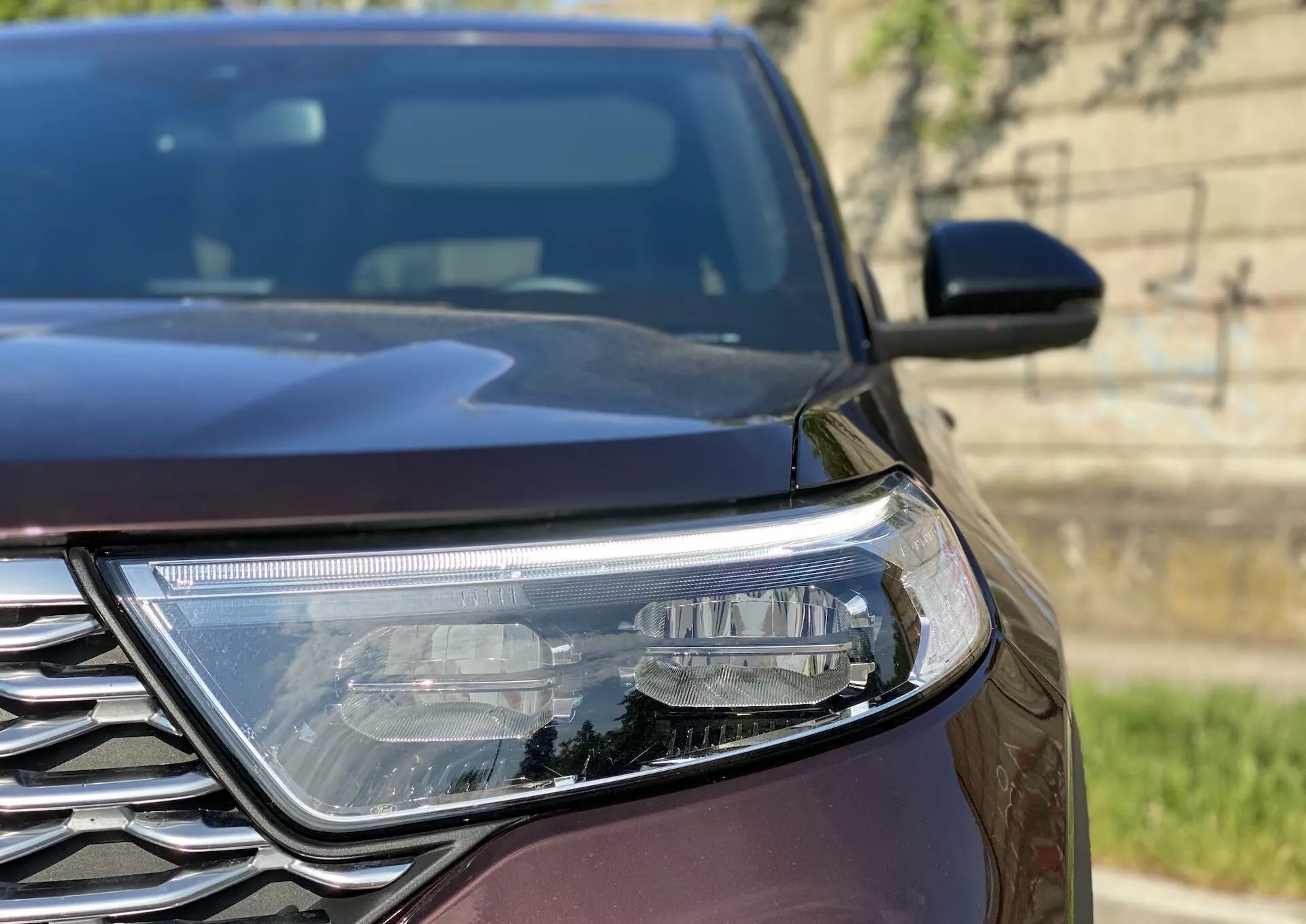 Zbliżony, skośny widok skupiający się na lewym reflektorze i części grilla Forda Explorera 2019, podkreślający kanciaste linie projektowe reflektora LED oraz złożony wzór chromowanej kratki w naturalnym świetle dziennym.