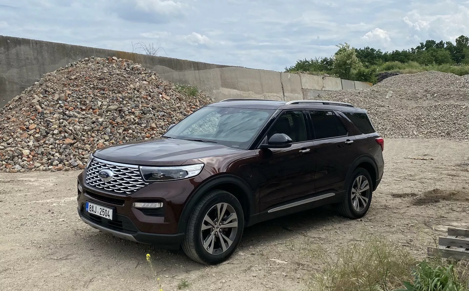Obraz przedstawia Ford Explorer 2019, nowoczesne SUV, zaparkowane na nierównym terenie przypominającym plac budowy lub teren off-road. Uchwycony z perspektywy trzech czwartych, zdjęcie pokazuje przód i bok pojazdu, w tym kratkę, reflektory, przedni zderzak, maskę, aluminiowe felgi, przednie i tylne drzwi, lusterka boczne z zintegrowanymi kierunkowskazami, błotnik i okna. Samochód jest skierowany w prawo, a metaliczne wykończenie lakieru jest wyraźnie widoczne, zapewniając pełny widok tego zadbanego pojazdu z relingami dachowymi sugerującymi dodatkową pojemność ładunkową.