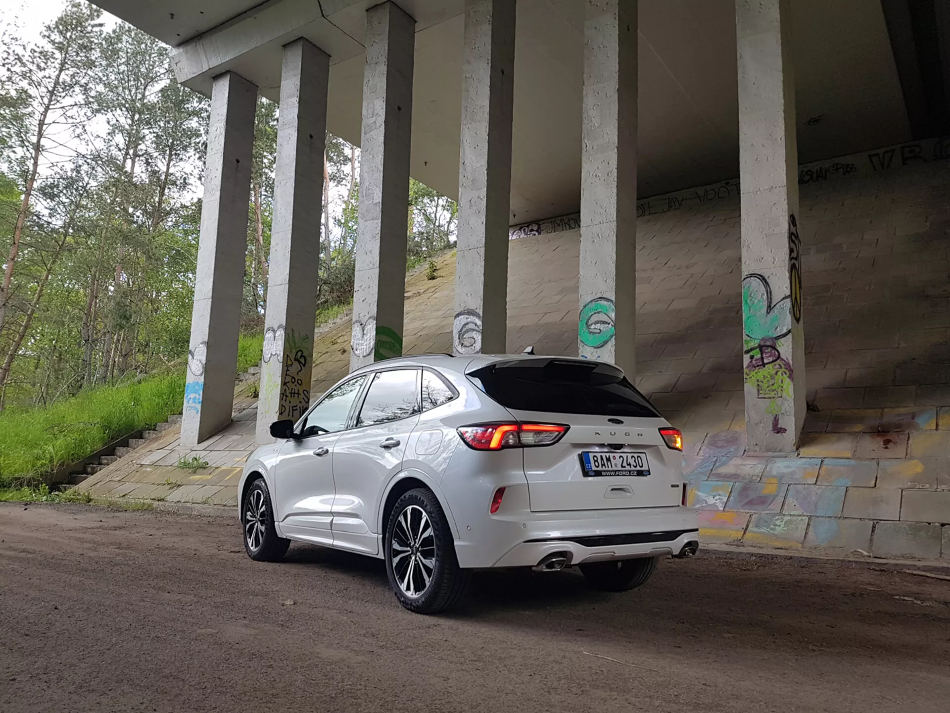 Crossover SUV Ford Kuga 2019 zaparkowany pod wiaduktem, prezentujący swój nowoczesny design i cechy technologiczne, takie jak diody LED w tylnych światłach, podwójny wydech i kamera cofania, wśród miejskich graffiti i betonowych filarów