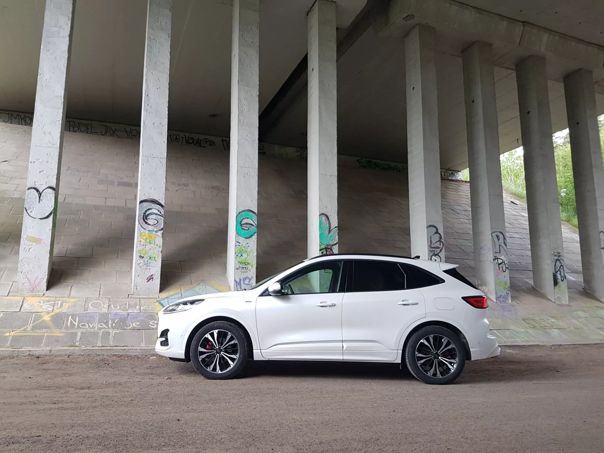 Biały kompaktowy SUV Ford Kuga z 2019 roku zaparkowany pod konstrukcją z betonowymi filarami pokrytymi graffiti, prezentujący profil przedniej ćwiartki, w tym grill, reflektory i koła ze stopu