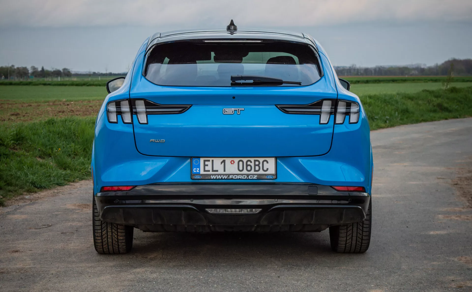 Tylny widok niebieskiego Forda Mustang-Mach-E z 2020 roku na asfaltowej drodze z polami trawy w tle, prezentujący kluczowe cechy takie jak logo producenta, nazwa modelu, emblemat wersji, polska tablica rejestracyjna, podwójny układ wydechowy, nowoczesne tylne światła LED, czujniki parkowania i wysoko zamontowane światło stop.