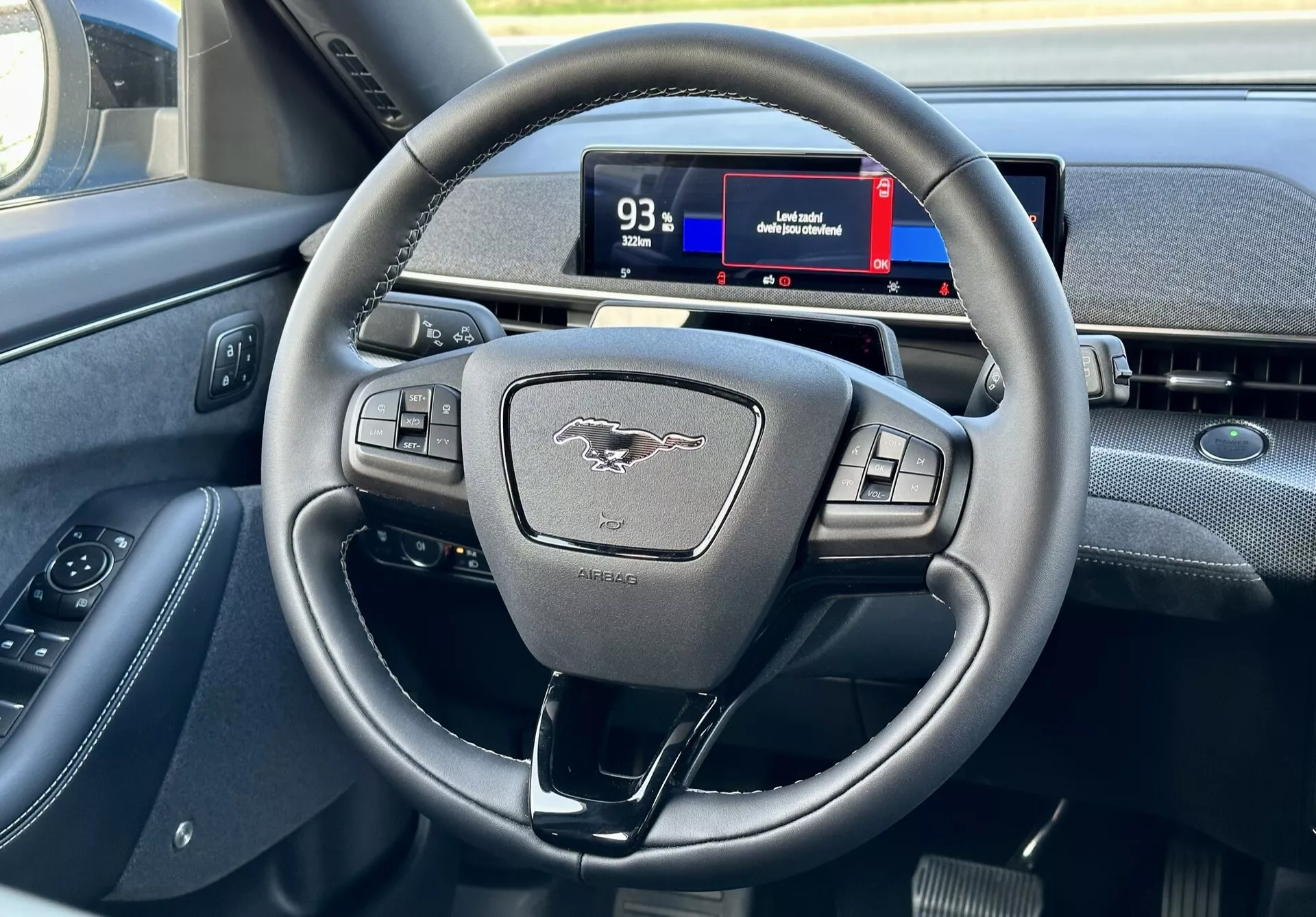 Wnętrze Ford Mustang Mach-E 2020 podkreślające nowoczesną kierownicę trójramienną z multifunkcyjnymi przyciskami i cyfrowym zegarem z perspektywy kierowcy, pokazujące elementy sterujące, emblemat i wskazówki nawigacyjne.