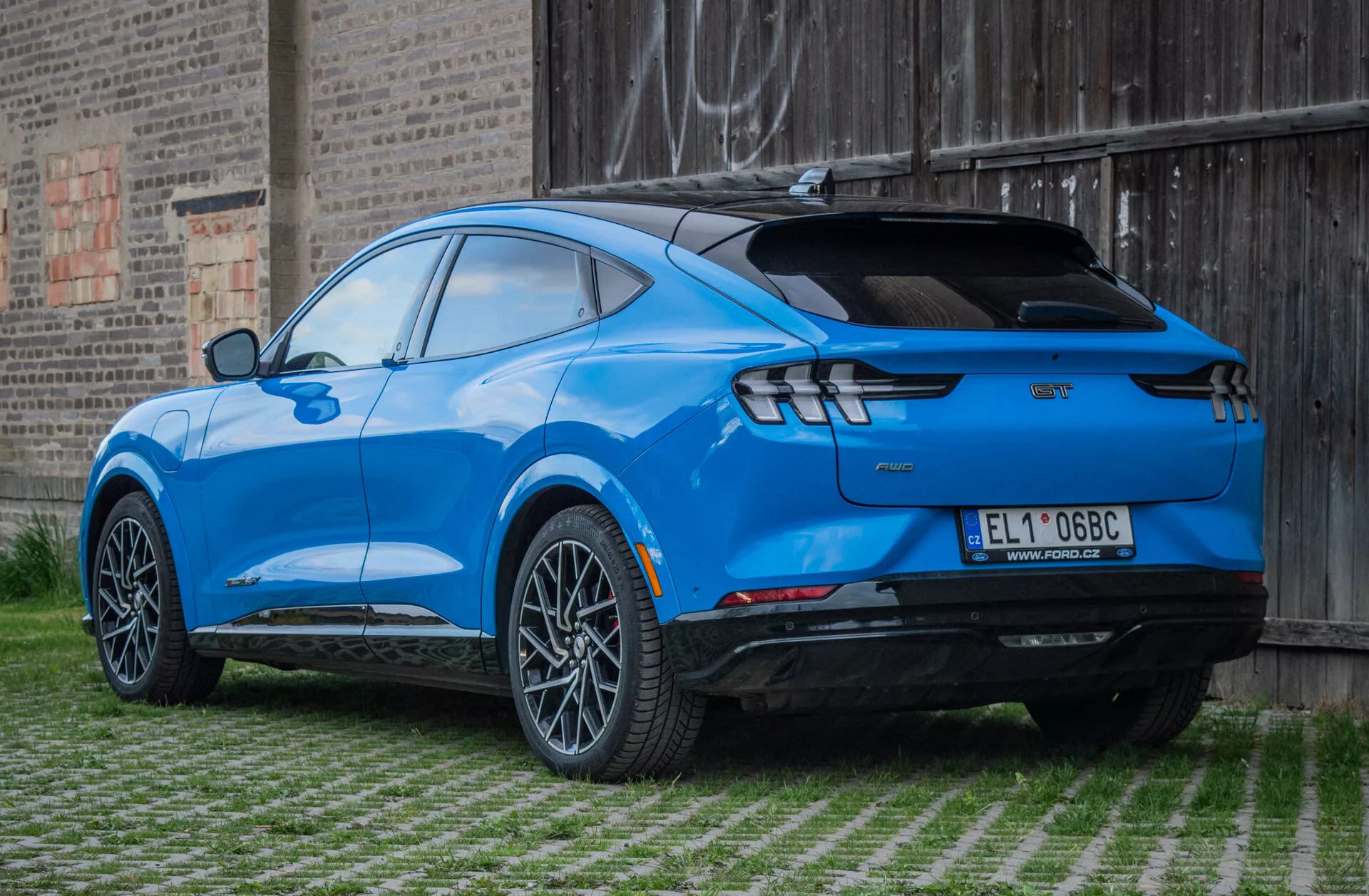 Ford Mustang Mach-E 2020 w jasnym niebieskim kolorze, widok z tyłu pod kątem trzech czwartych pokazujący elegancki design, felgi ze stopów, ledowe tylne światła i sportowy spoiler, zaparkowany na obszarze z trawnikiem z tłem z masywu murowanego